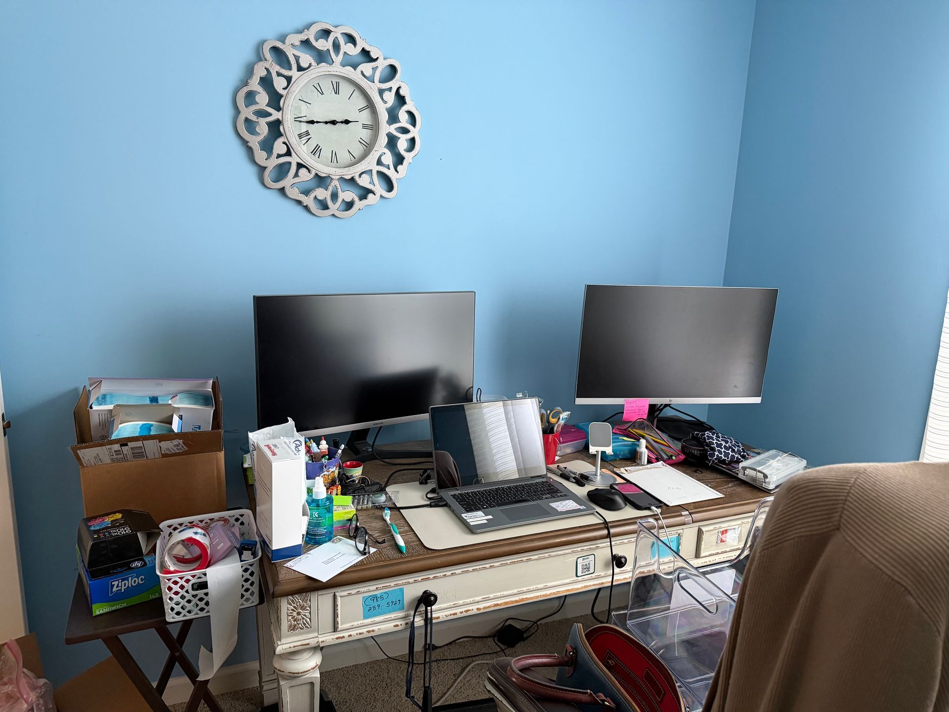 Home office desk with dual monitors against a blue wall and decorative clock above.