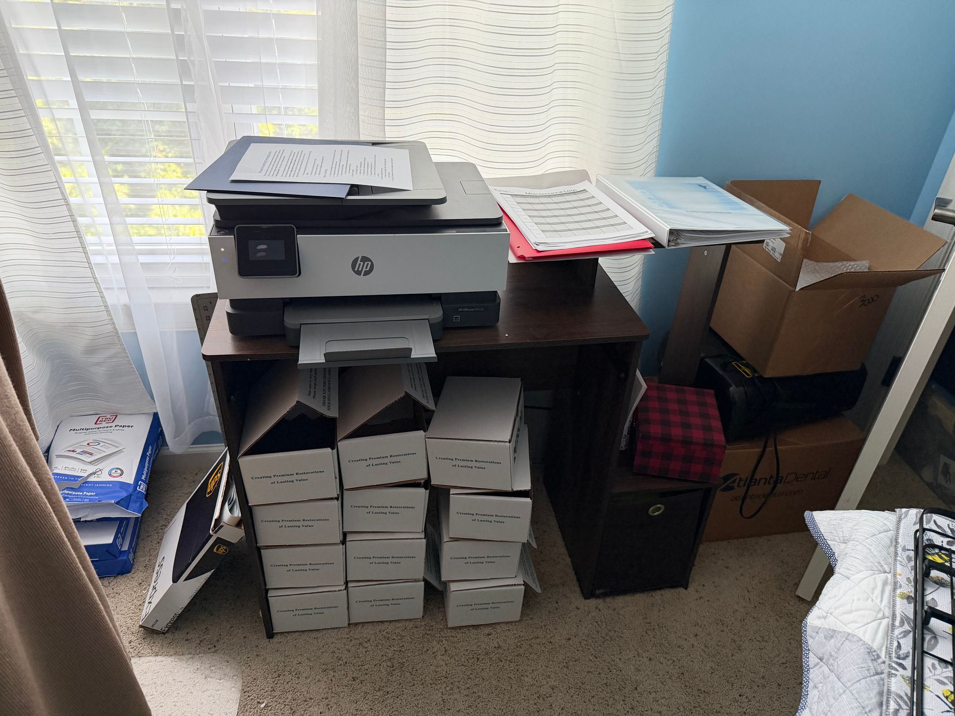 Cluttered office corner with printer on a shelf, stacked papers, and boxes by the window.