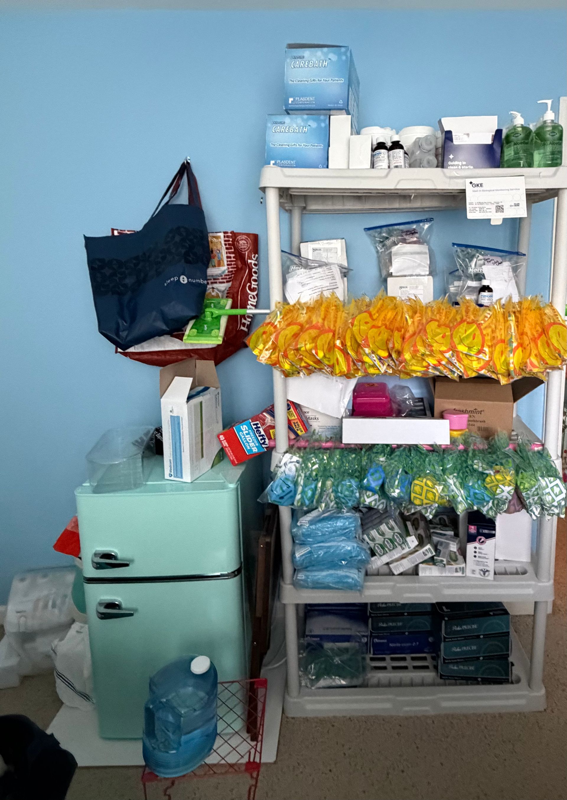 Shelf of assorted household goods and snacks against a blue wall, with a small fridge and water jug nearby