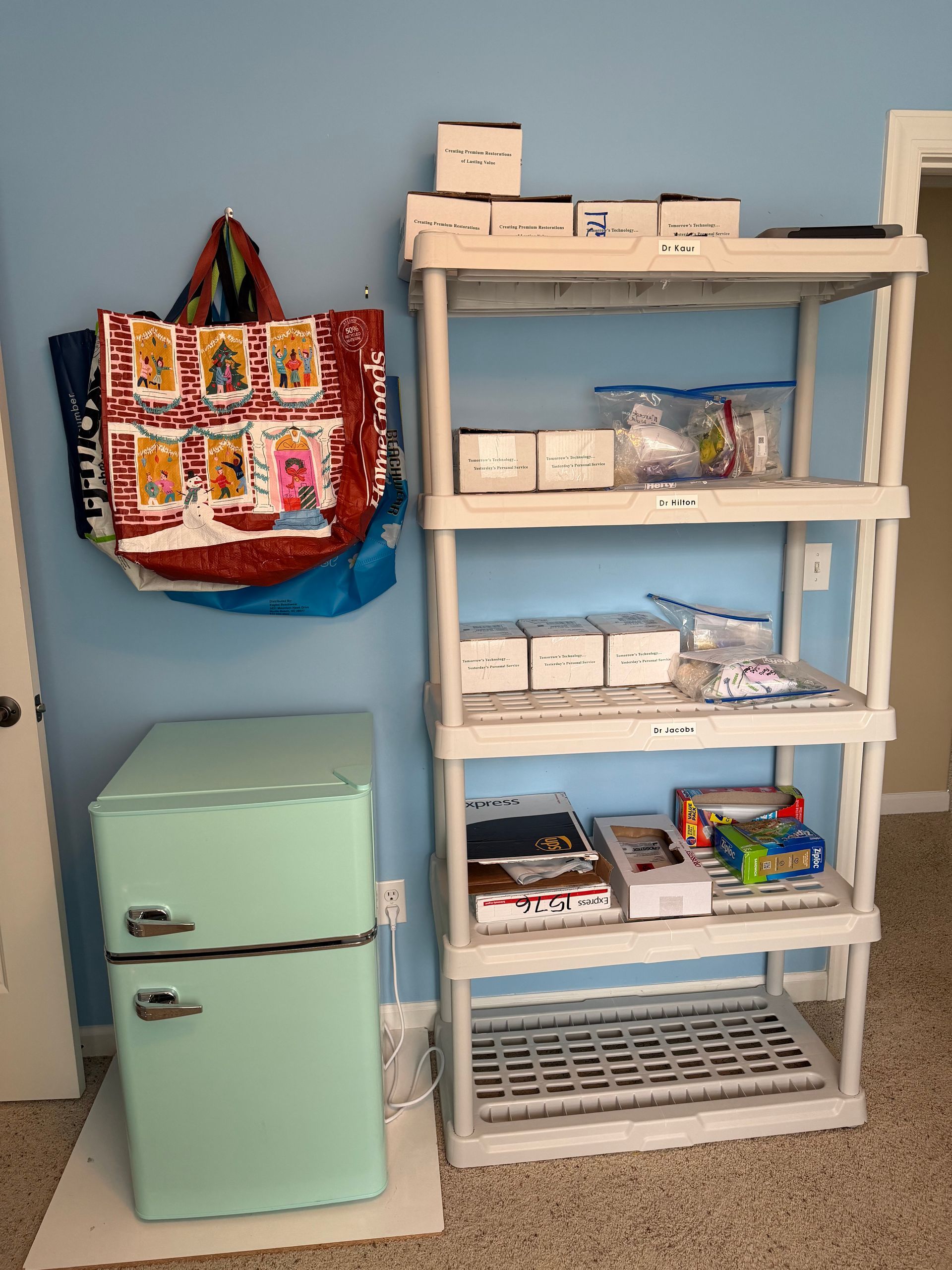 Mint mini fridge beside white shelving with boxes, baskets, and supplies against a blue wall.