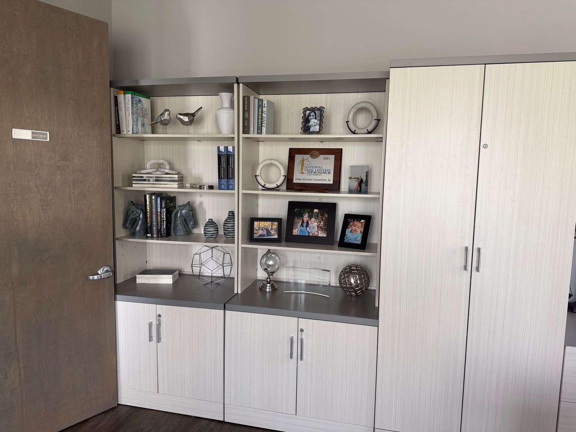 Office shelving with framed photos, trophies, and white cabinets beside a wooden door