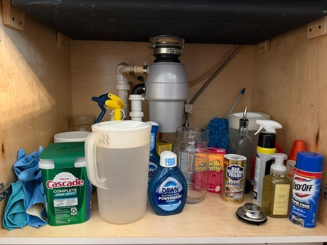 Under-sink cabinet with assorted cleaning supplies, bottles, and a container on a wooden shelf