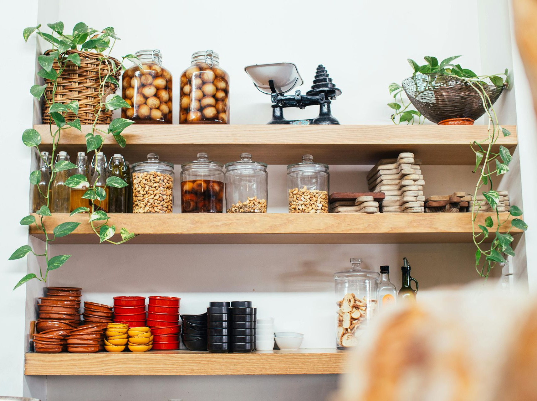 Wooden shelves with jars of dry goods, potted plants, and a small vintage scale in a bright kitchen