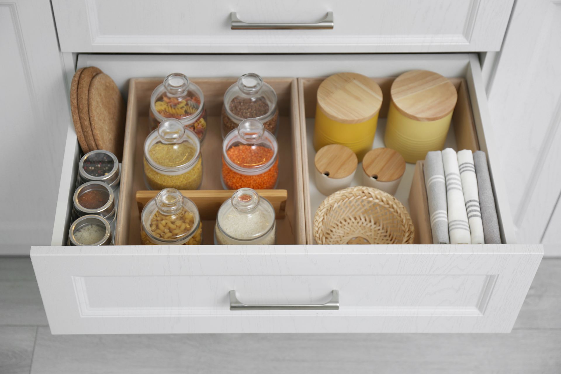 Open kitchen drawer organized with glass jars, candles, napkins, and woven baskets