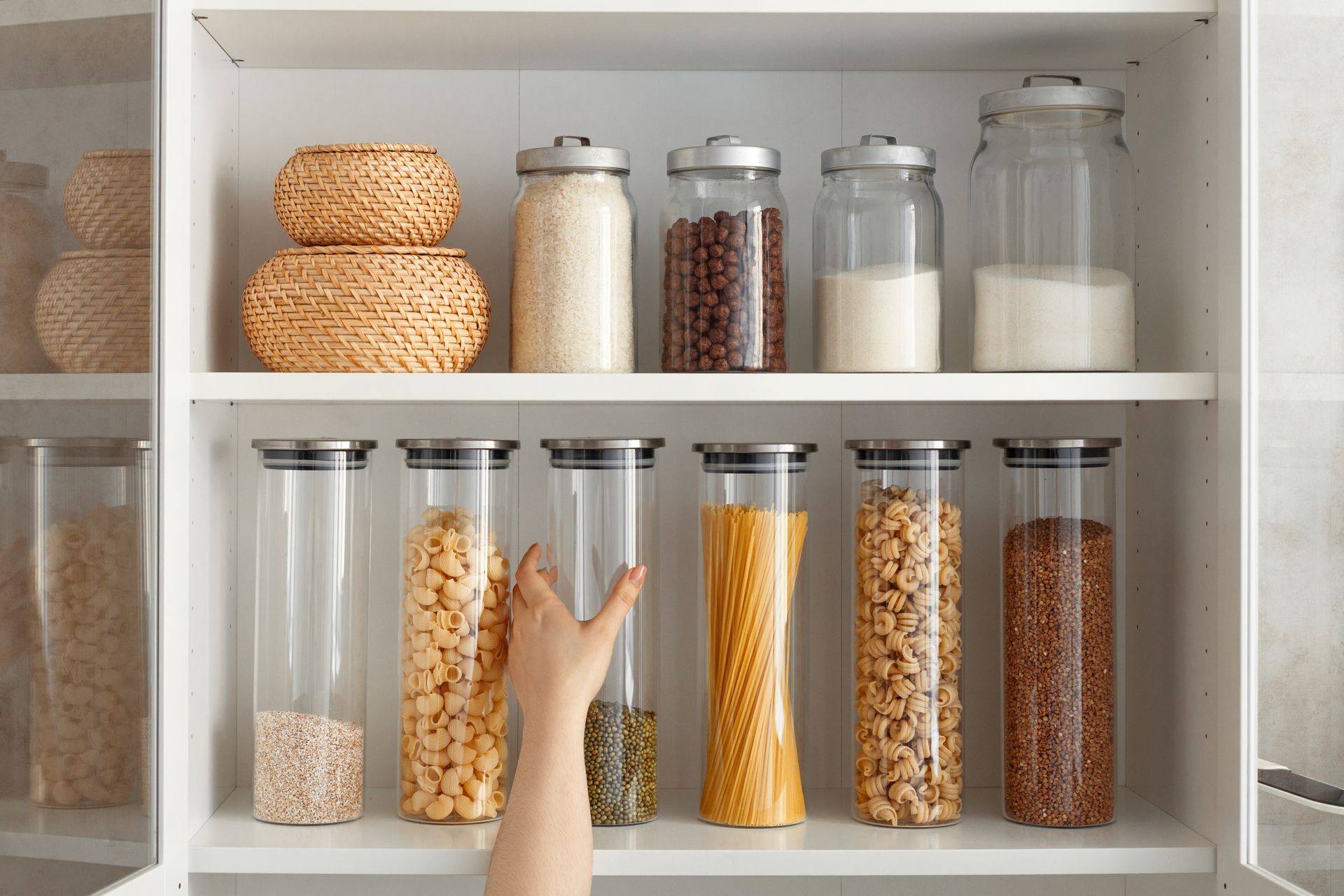 Open pantry shelves with glass jars of dry goods and stacked bread, with a hand reaching toward a middle jar.