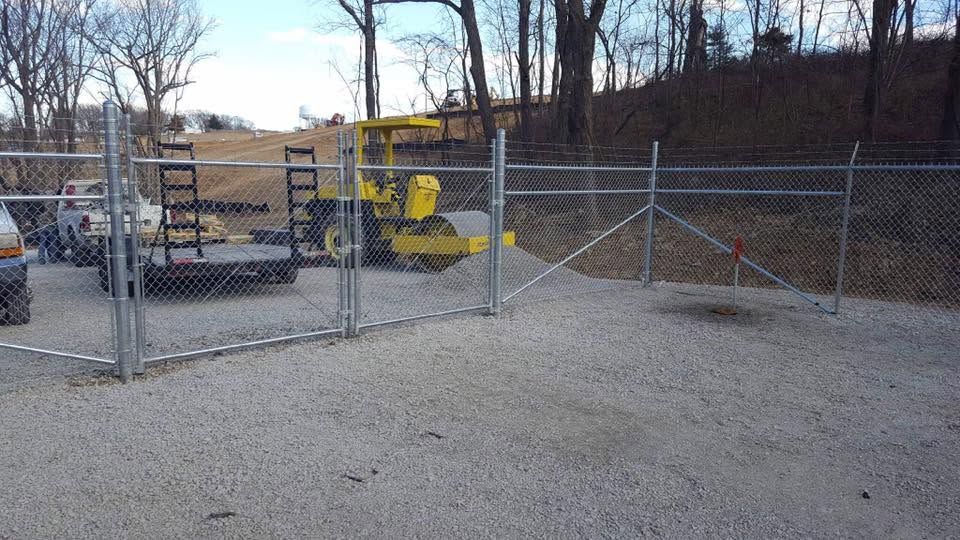 Chain-link gate at a gravel lot with a yellow utility machine and leafless trees in the background