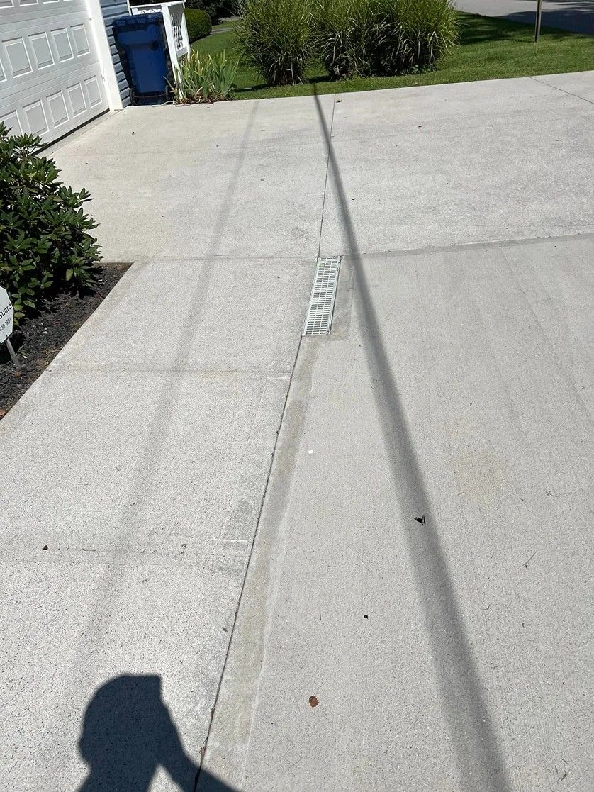 Concrete driveway with a narrow drainage channel, grass and bushes at the edge, and a shadow in the foreground