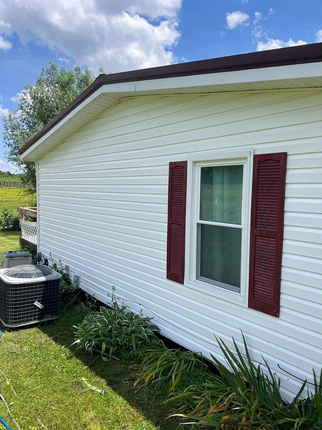 White house siding with maroon shutters and a window beside a lawn under a blue sky.