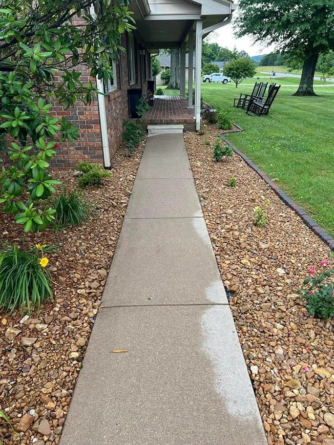 Front walkway to a brick house, bordered by mulch and shrubs, with a wet concrete path and green lawn.