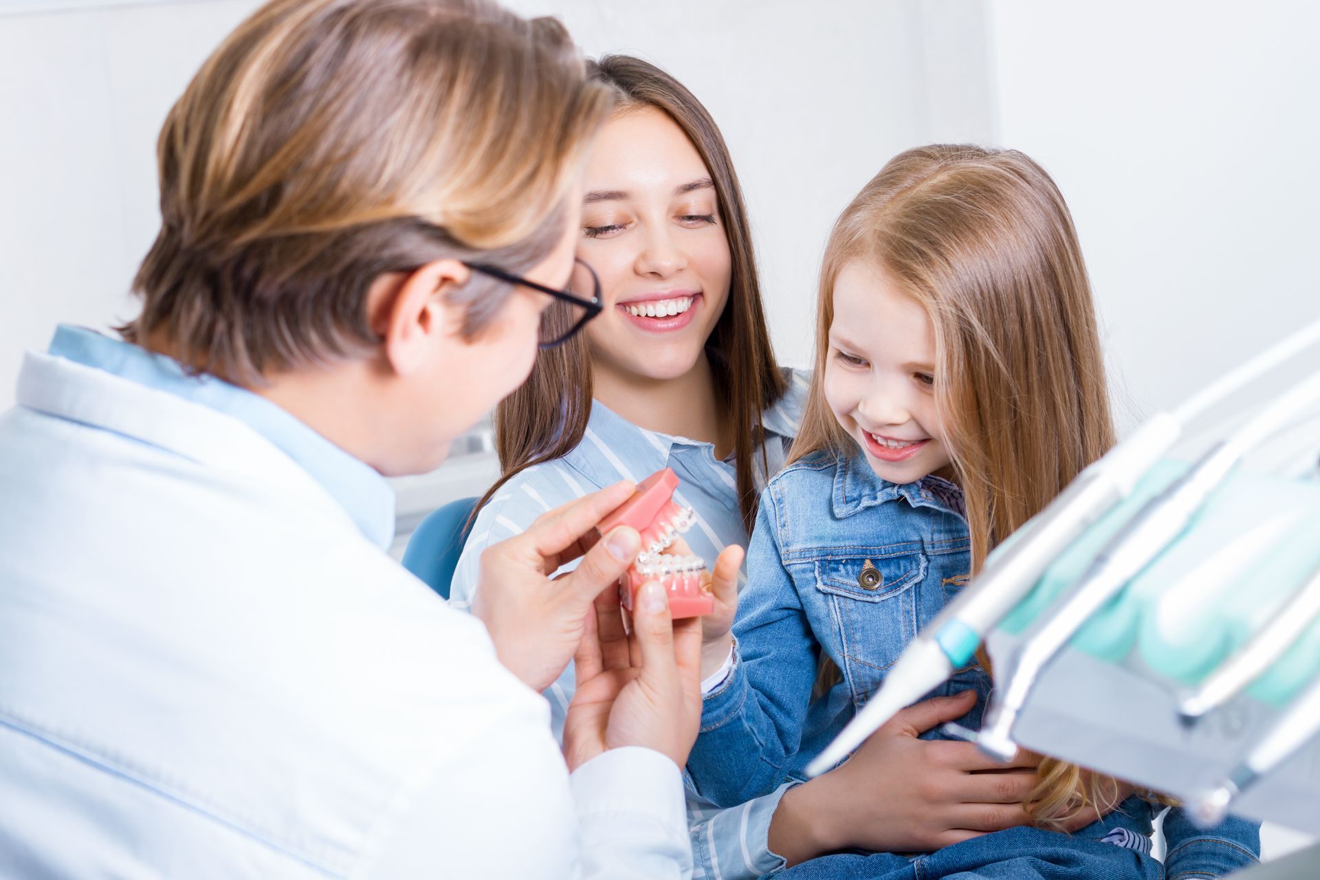 A girl is sitting in a dental chair with her mother while a dentist is showing them model teeth.
