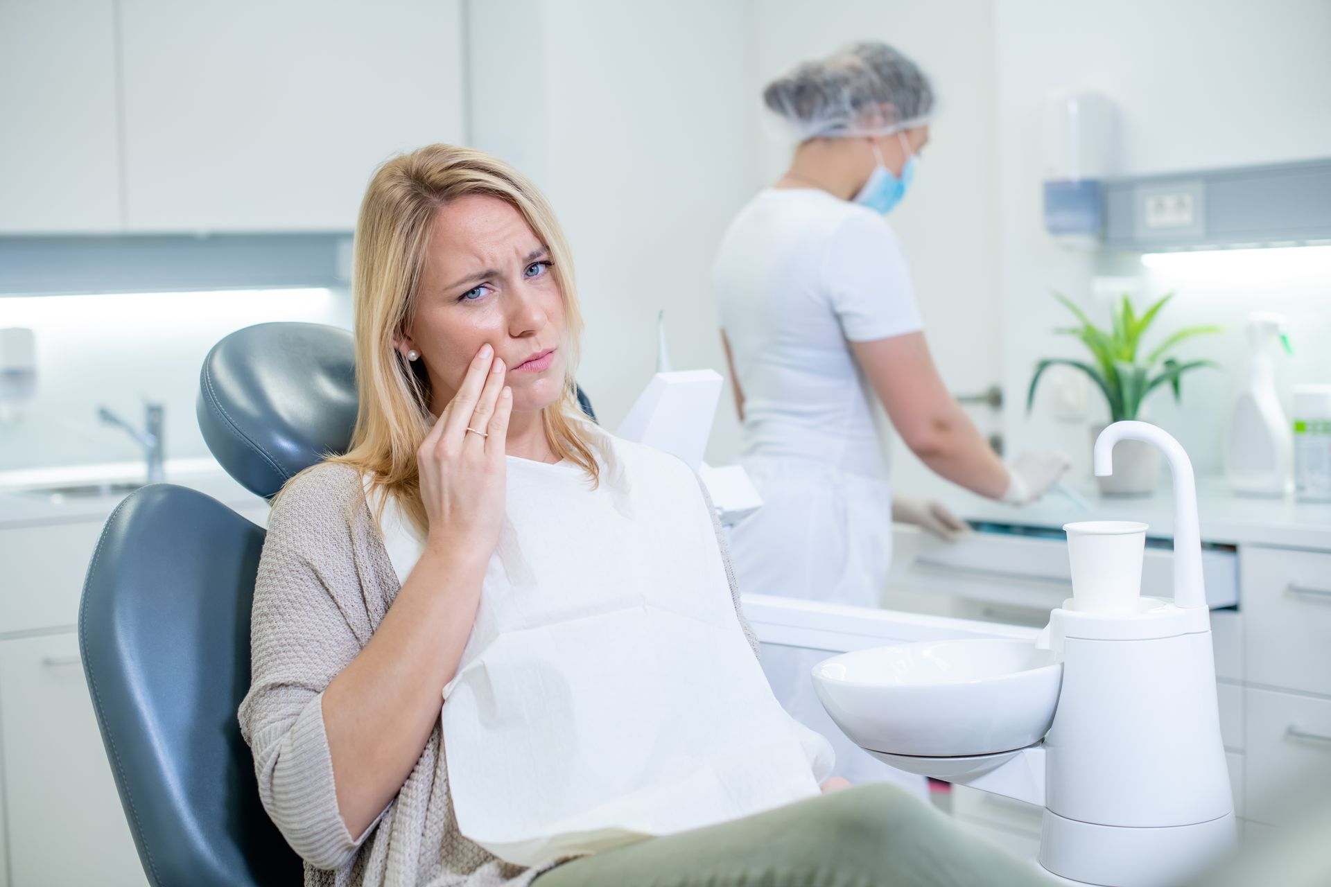 A woman suffering from a toothache while a dentist is preparing for examination in the background.