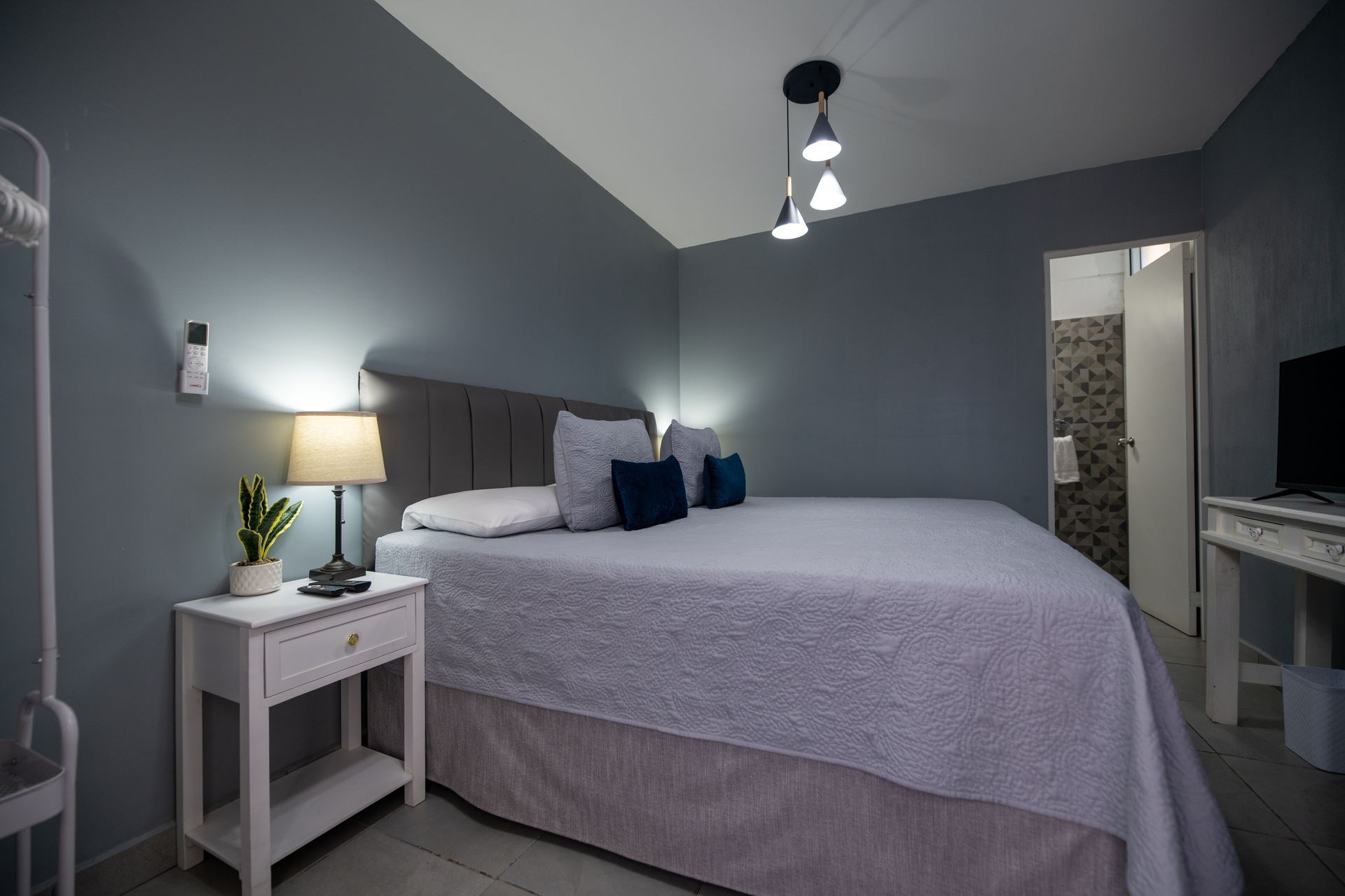 Habitación pequeña de color gris con una cama bien hecha, mesita de noche, lámpara y lámparas colgantes.