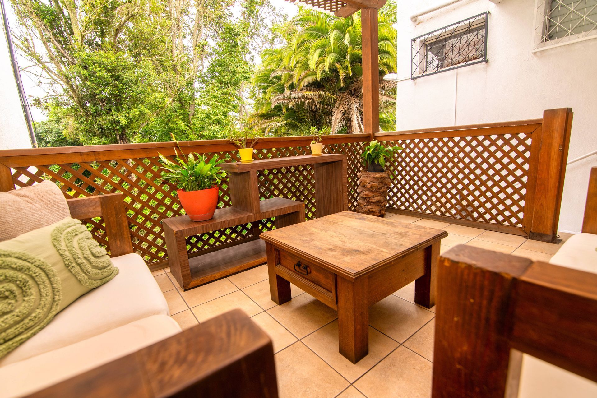 Acogedora zona de estar en el patio con cojines blancos, mesa de madera, barandilla de celosía y una planta en maceta.