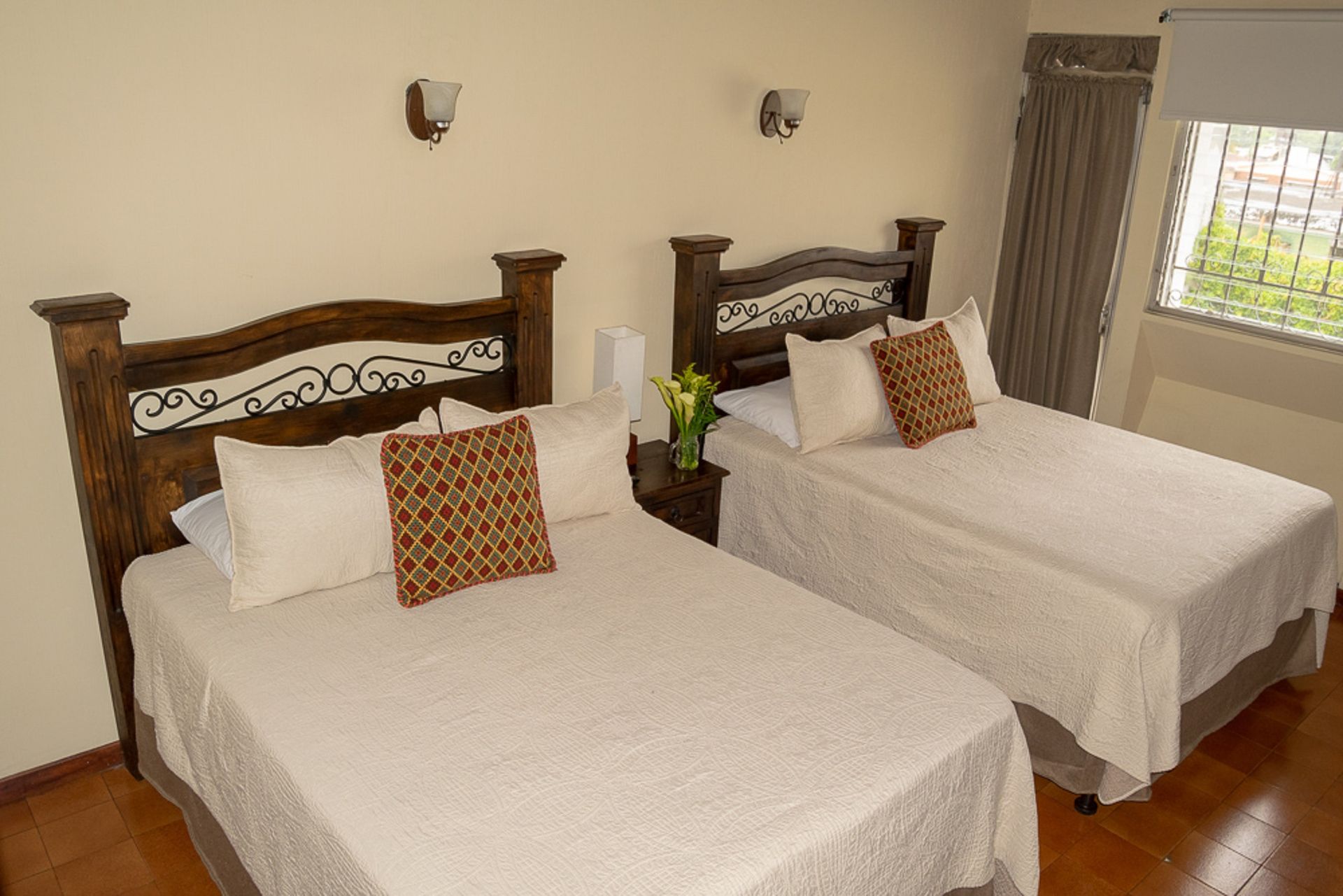 Habitación luminosa con dos camas individuales, colchas blancas, cabeceros de madera y una ventana con cortinas.