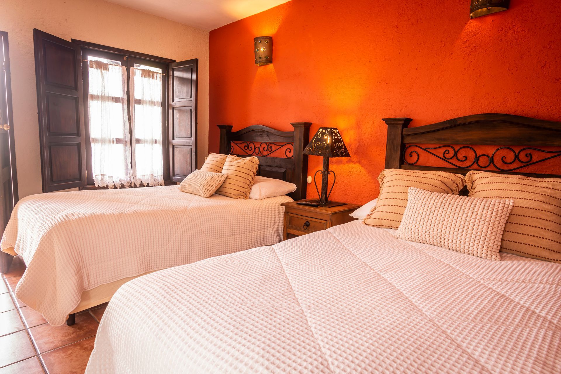 Habitación de color naranja cálido con dos camas blancas, almohadas estampadas y luz solar que entra por una ventana.