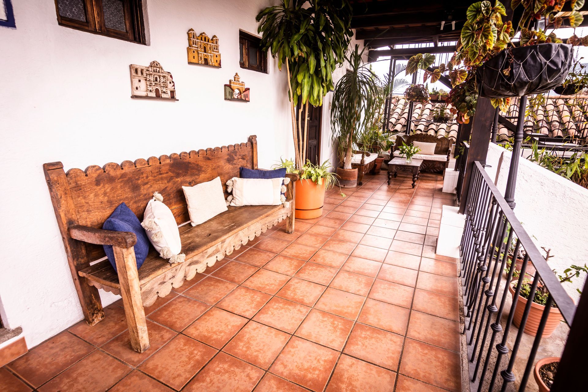 Porche con suelo de terracota, banco de madera, plantas colgantes y barandilla con vistas a la vegetación.