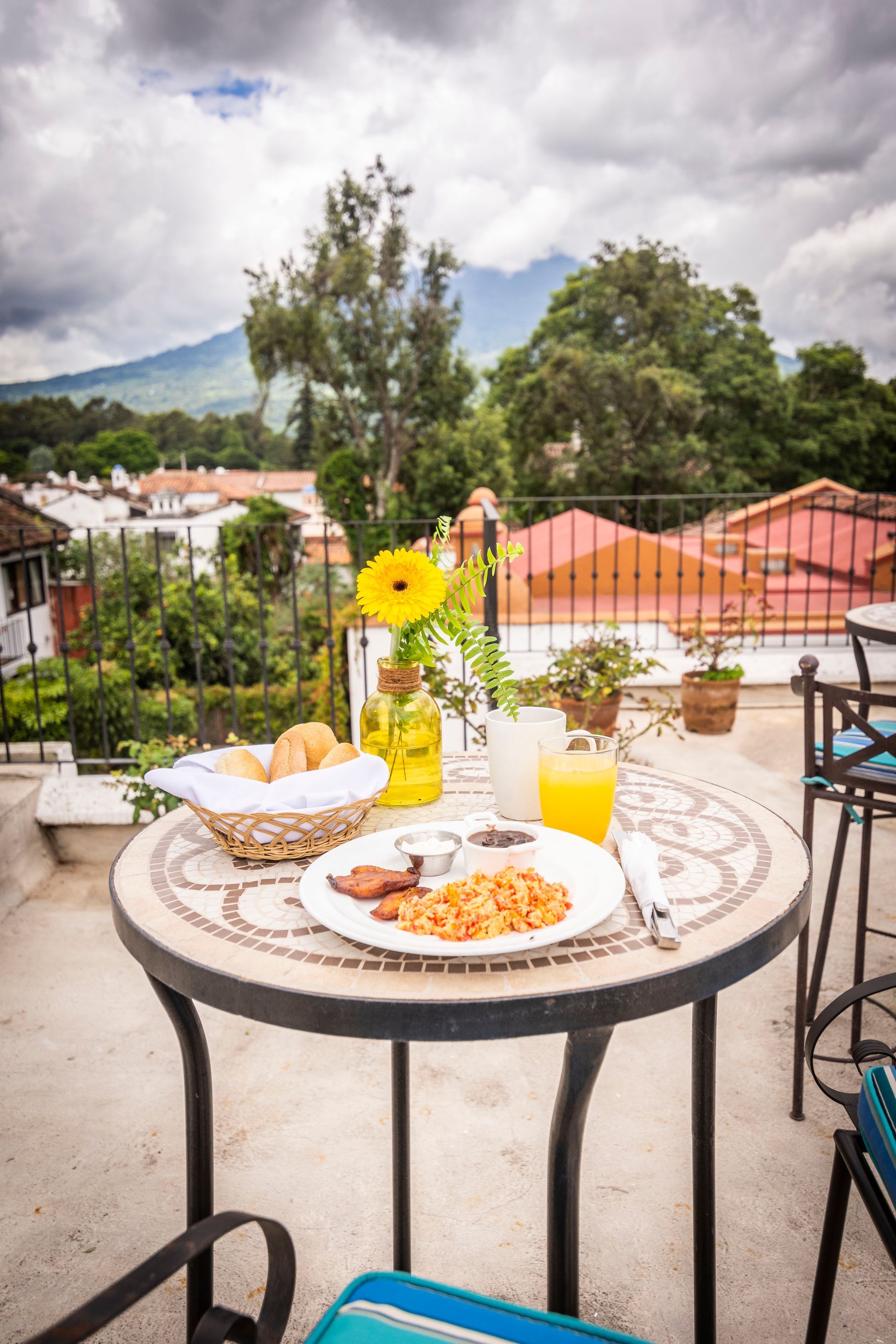 Mesa al aire libre con bollería, zumo de naranja y una flor amarilla, con vistas a tejados y montañas.