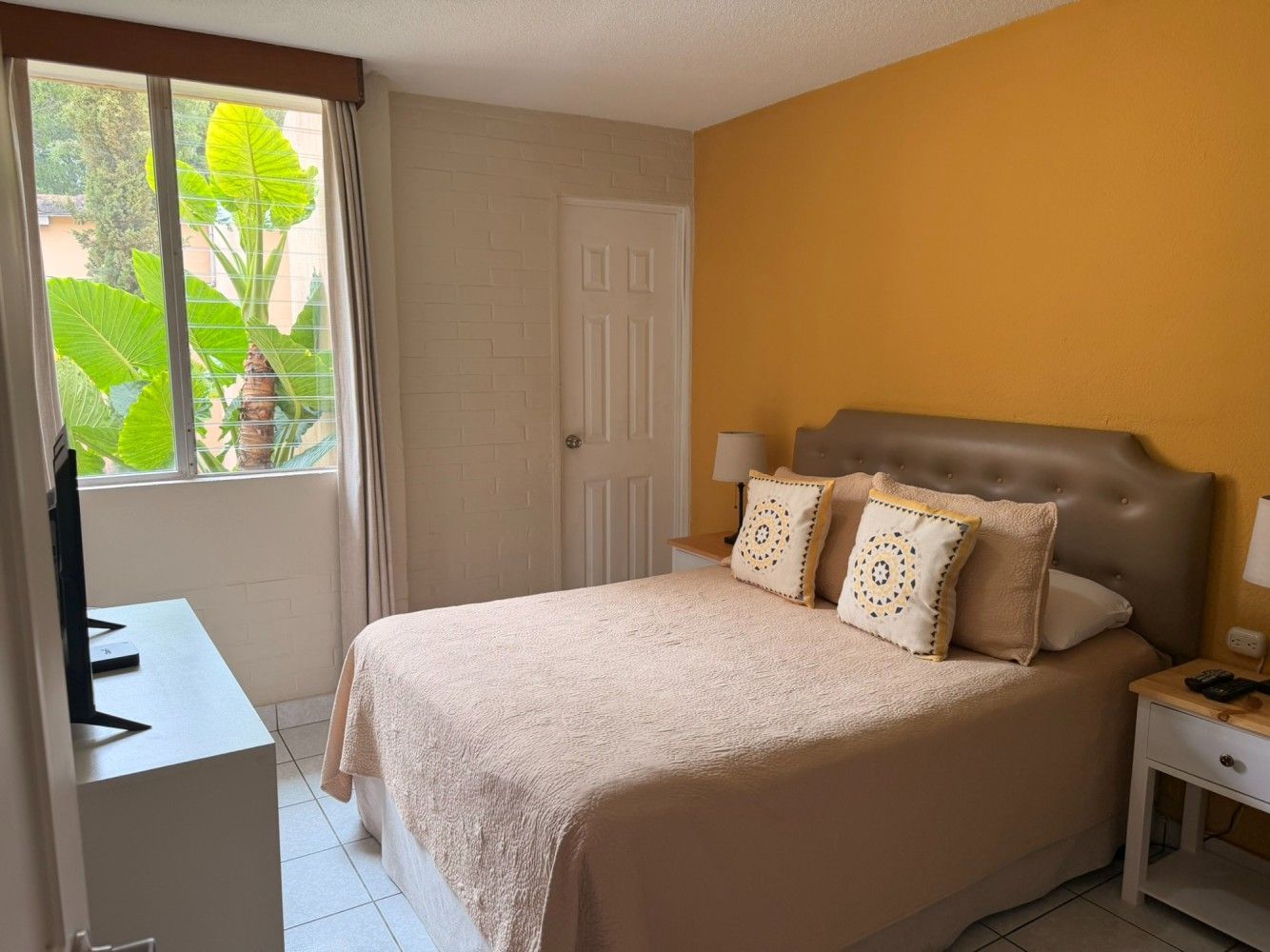 Habitación pequeña con cama, pared decorativa color mostaza y puerta con ventana que da a un balcón.