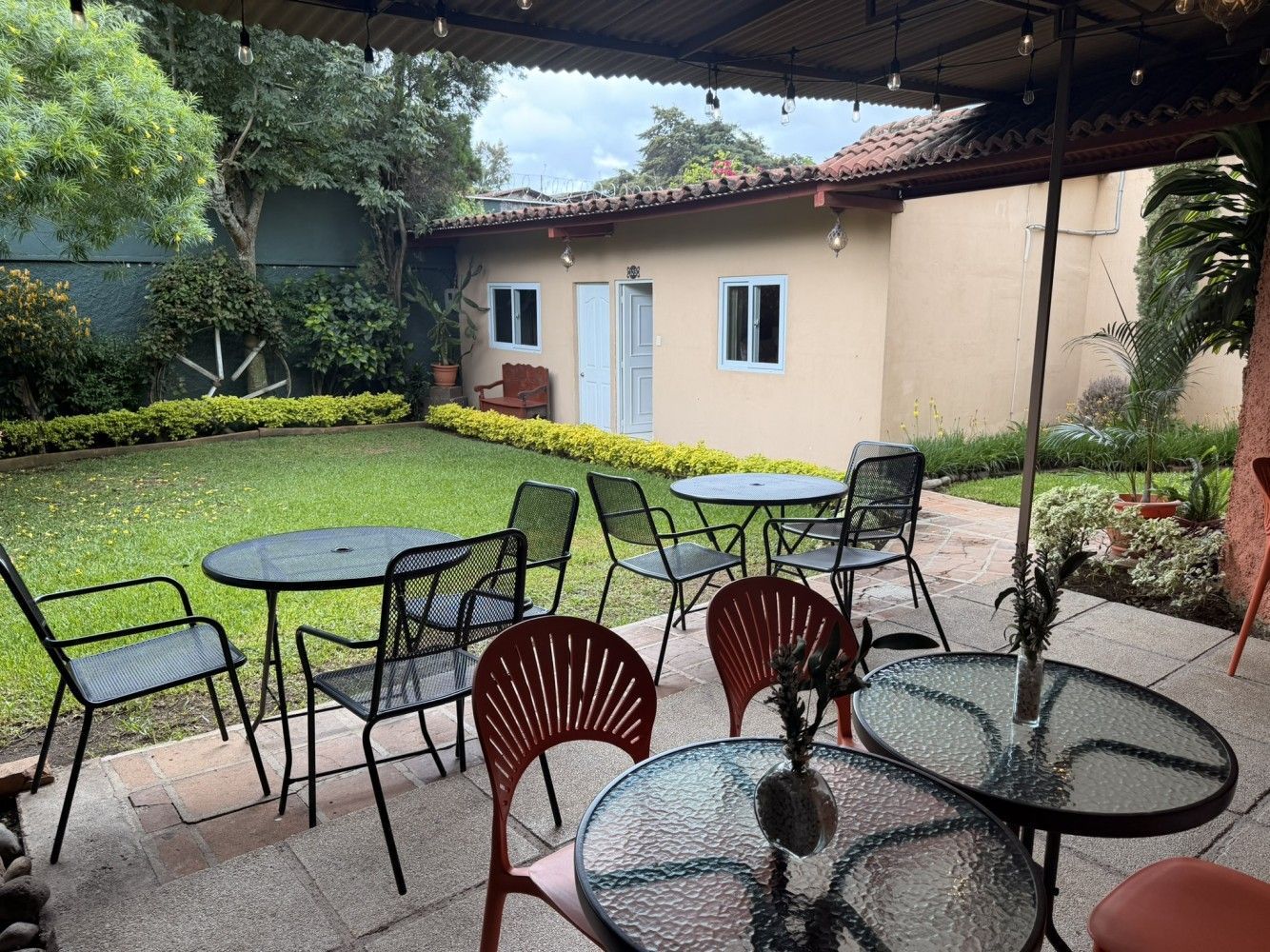 Patio con mesas de cristal y sillas de metal bajo un techo cubierto, junto a un pequeño edificio de color crema.