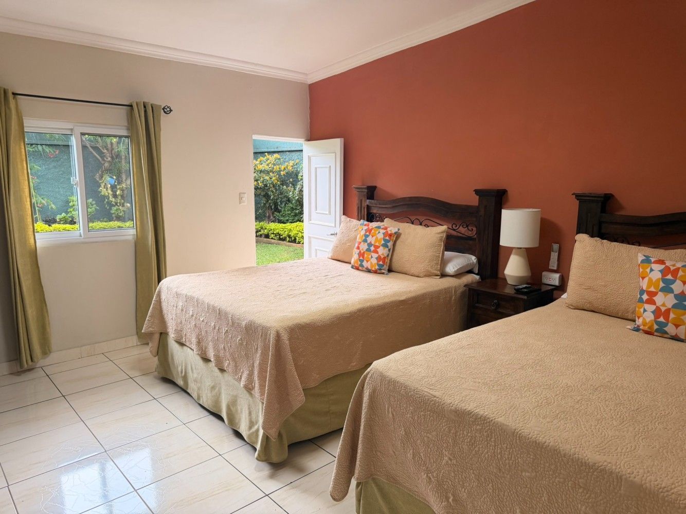 Habitación luminosa con dos camas, sábanas de color beige y una ventana con vistas a la vegetación y al océano.