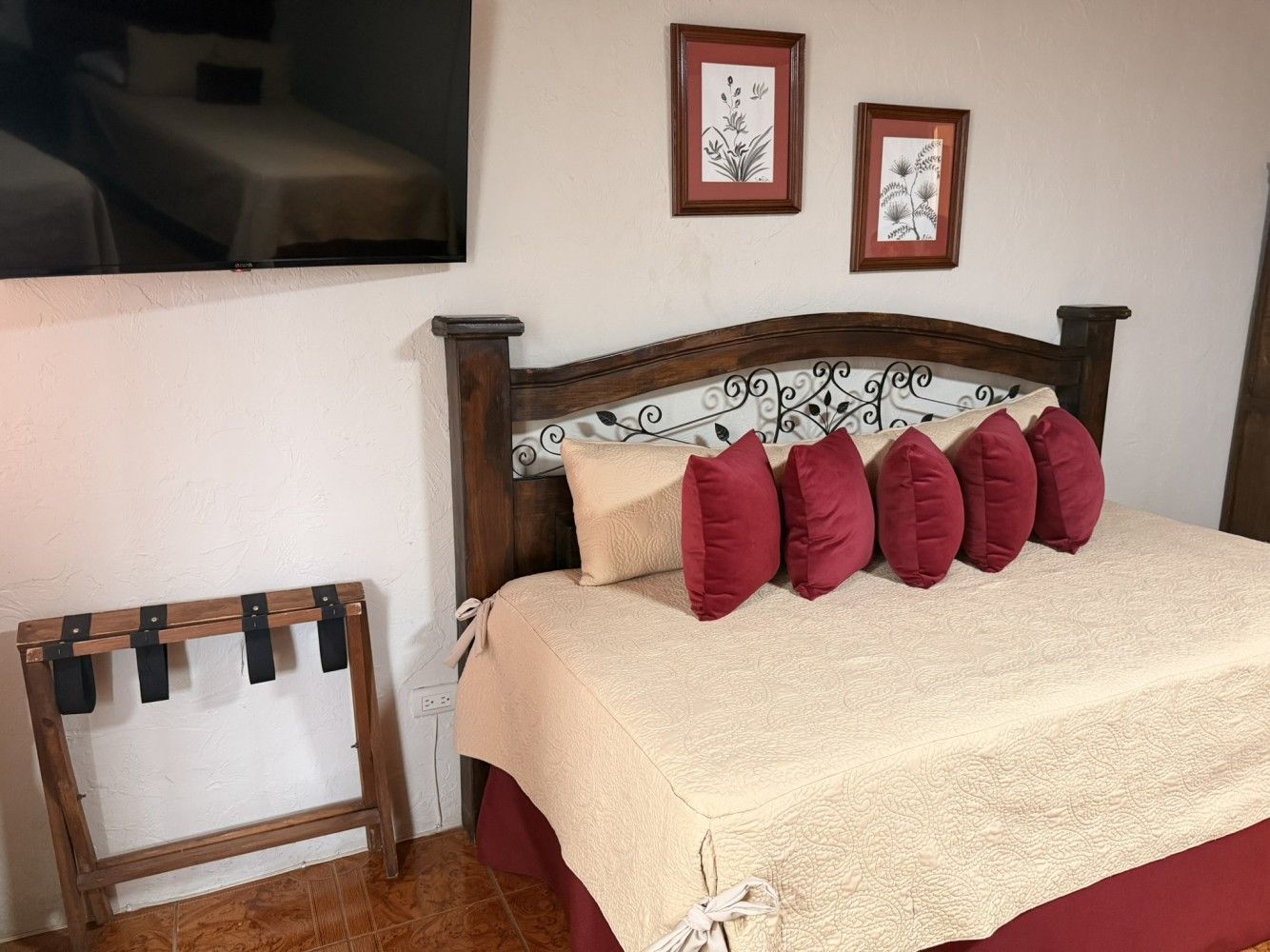 Dormitorio con cama de madera oscura, ropa de cama crema, almohadas rojas, cuadros decorativos y televisor.