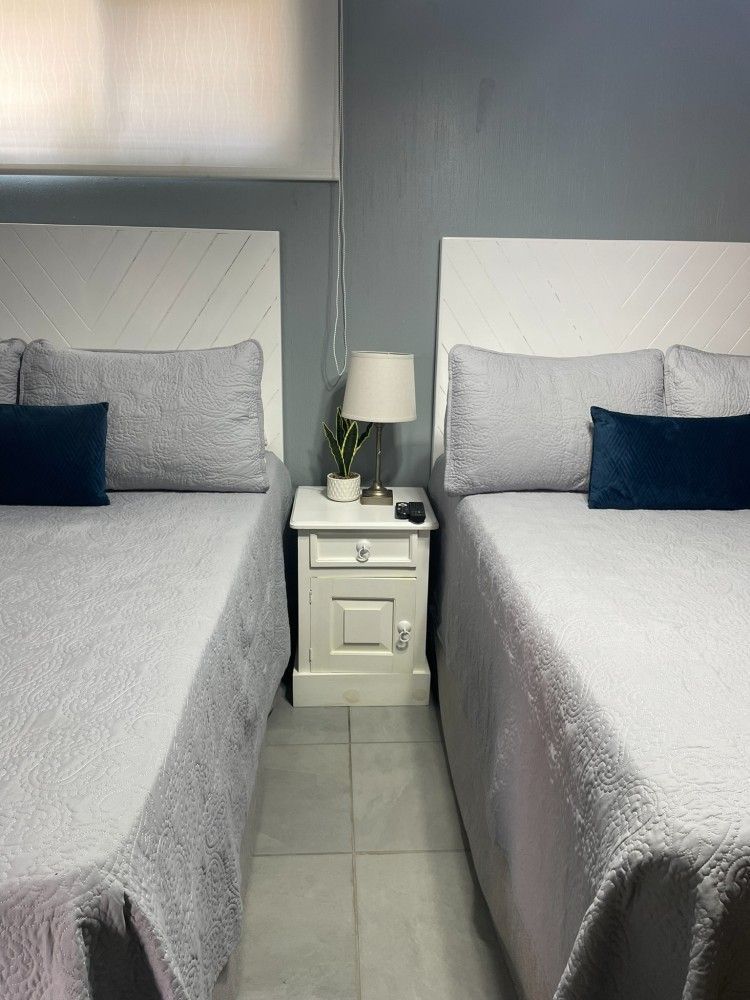 En una habitación gris, dos camas individuales con ropa de cama gris flanquean una mesita blanca con lámpara.