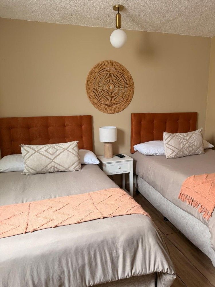 Dos camas individuales con cabeceros naranjas, mesita de noche, lámpara y decoración tejida.