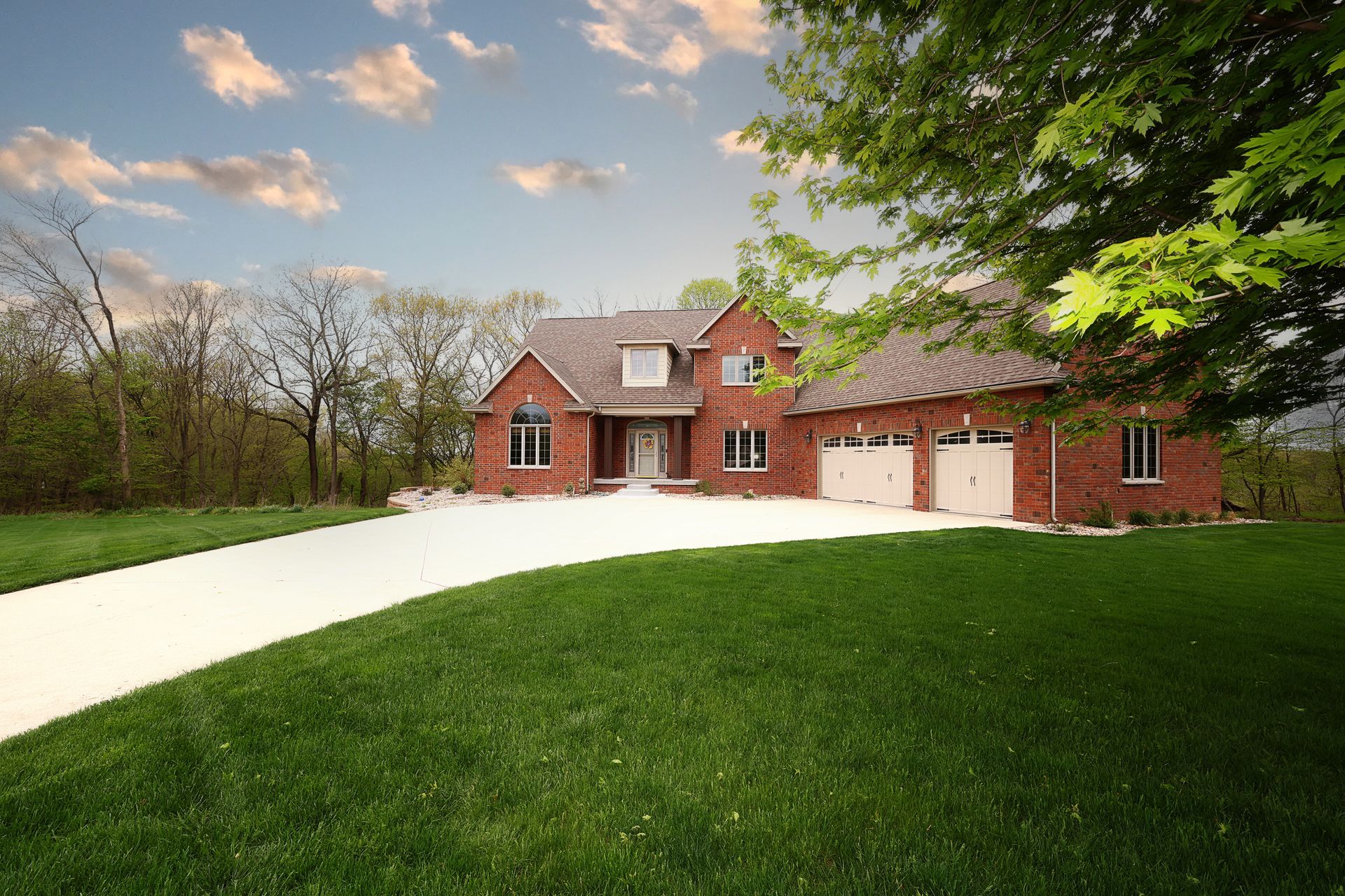Brick house with red shutters and garage beside a long white driveway and green lawn under a cloudy sky