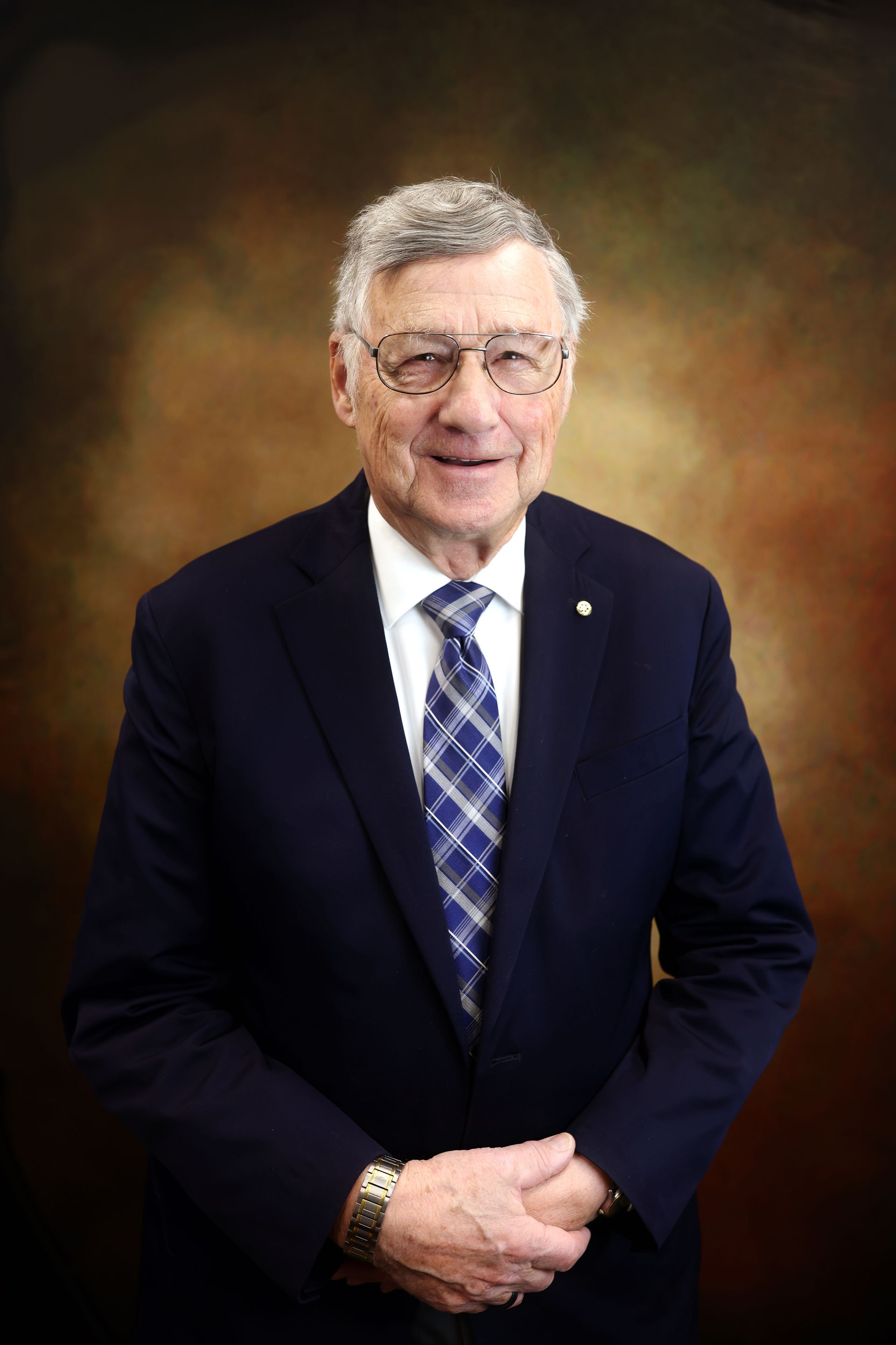 Studio portrait of a man in a dark suit and blue tie, standing with hands clasped against a warm background