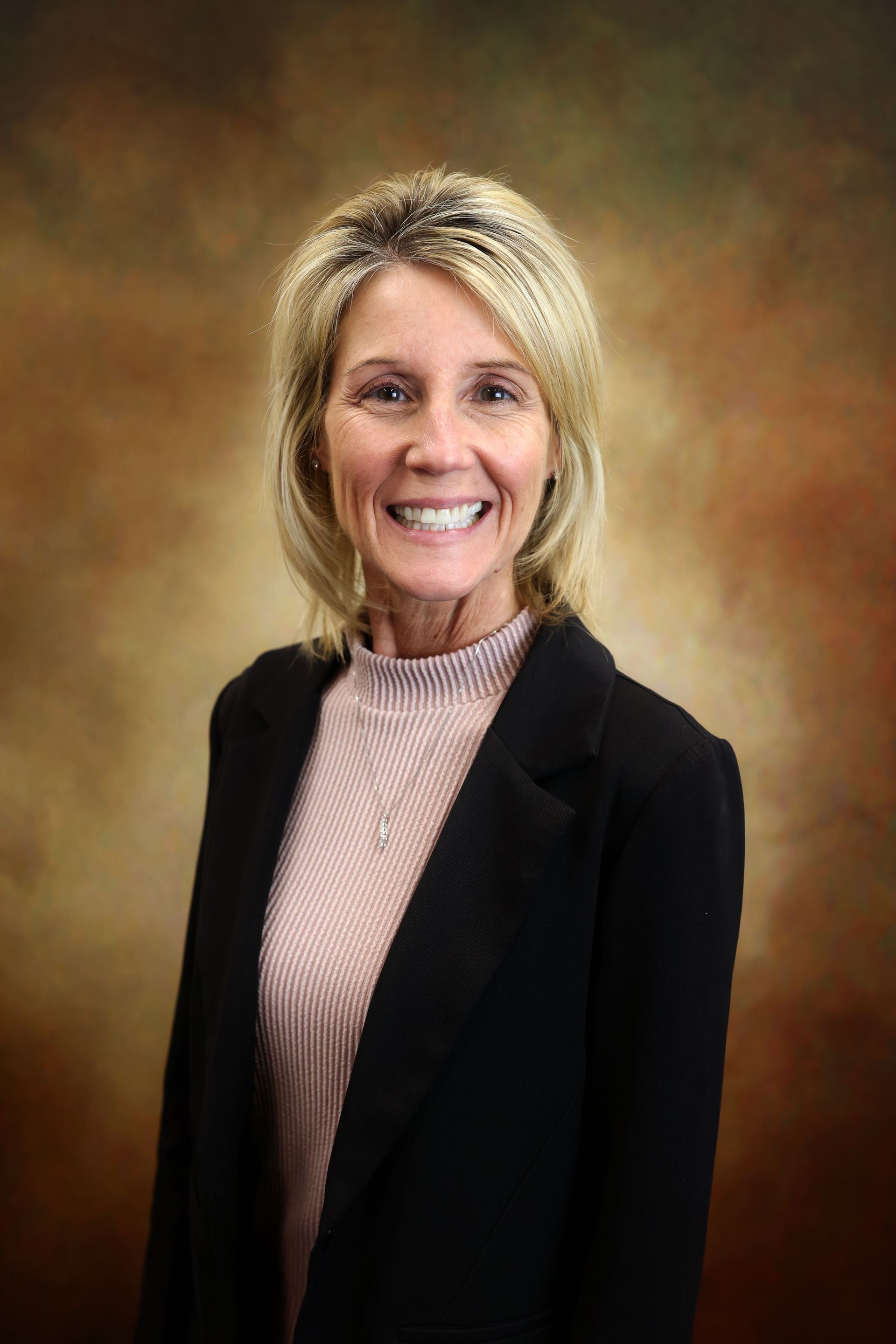 Smiling woman in a black blazer and light pink top against a warm studio backdrop