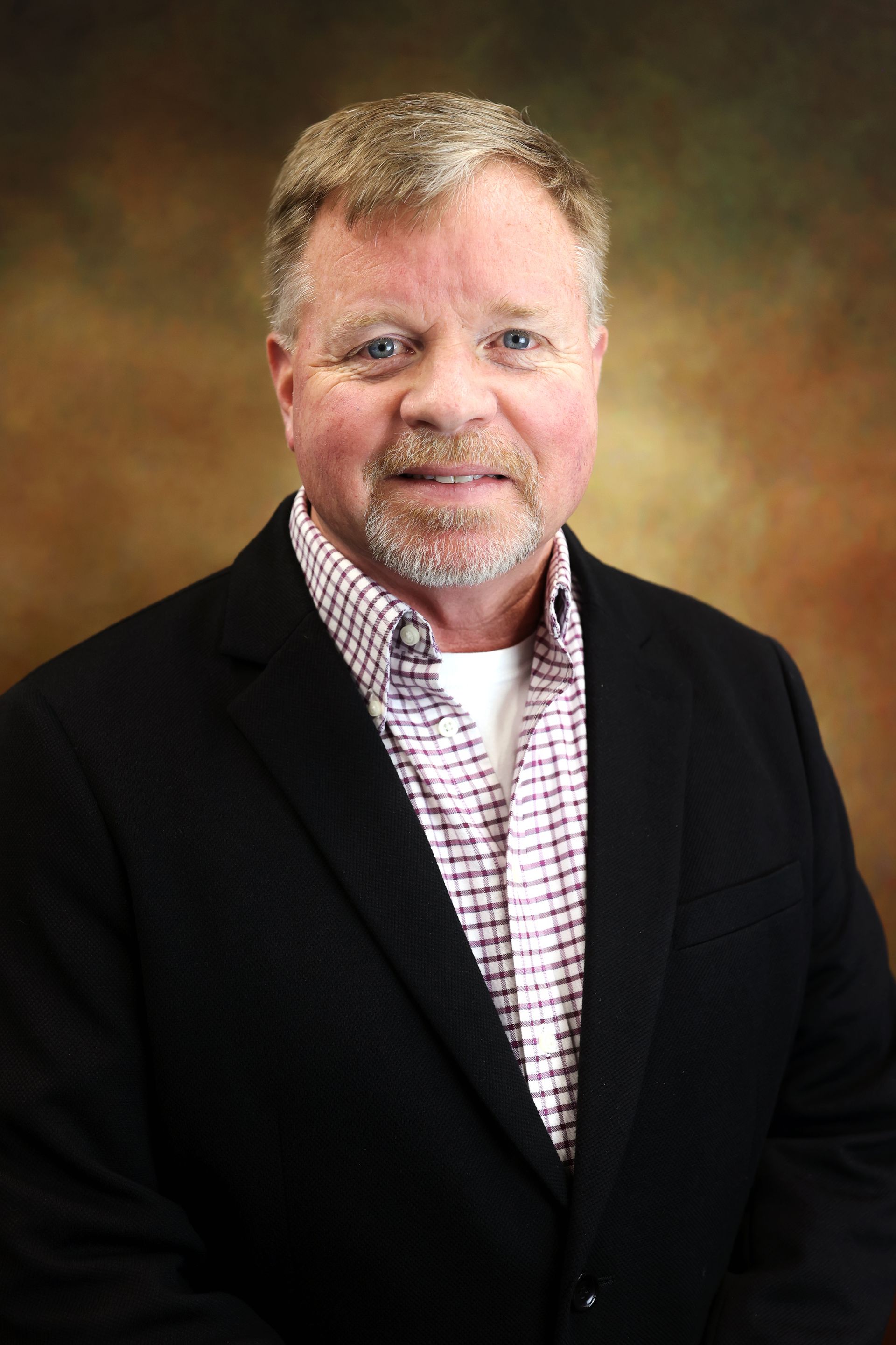 Smiling man in a black blazer and checkered shirt against a warm studio backdrop