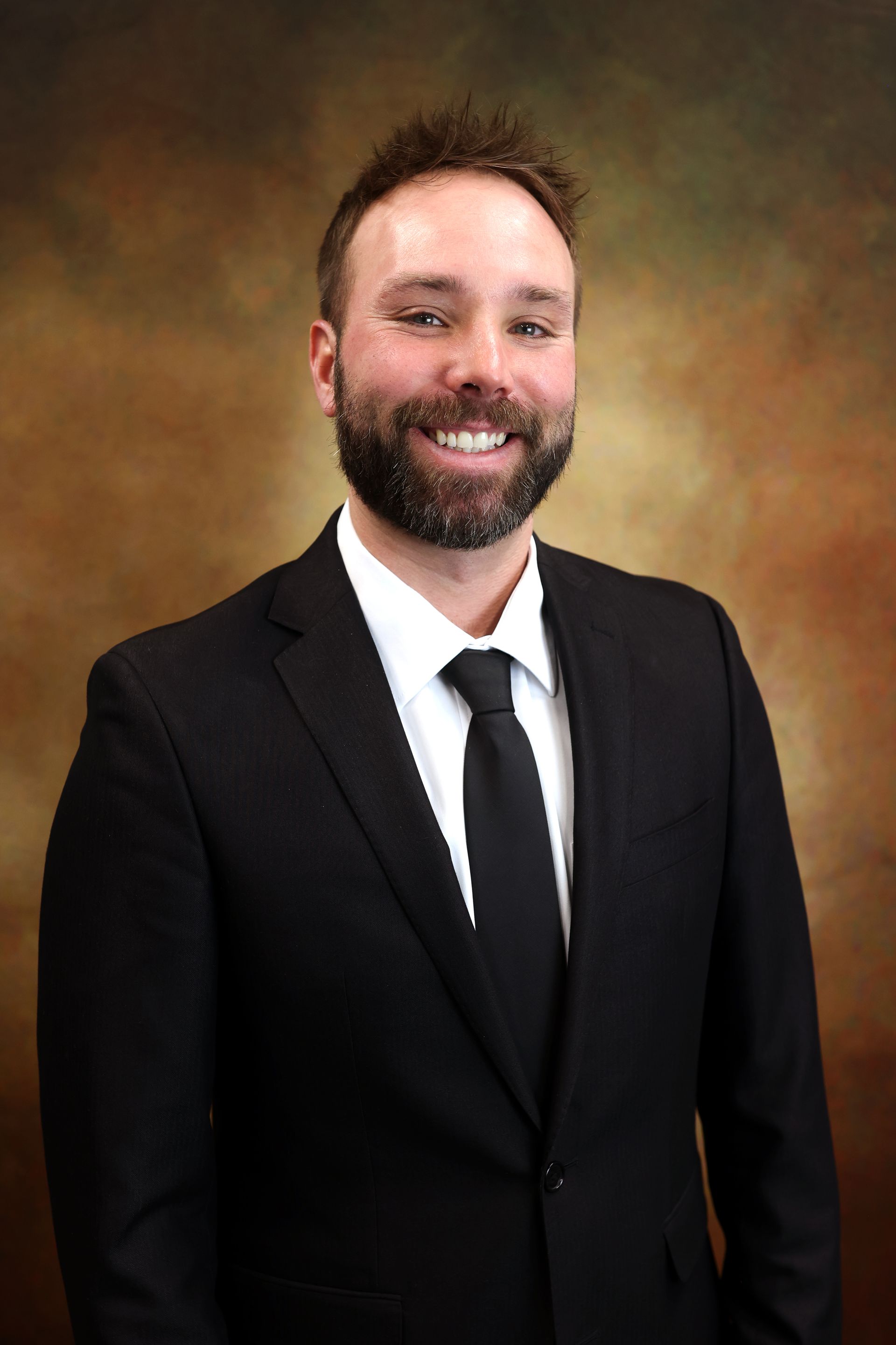 Smiling man in a black suit, white shirt, and black tie against a brown studio backdrop