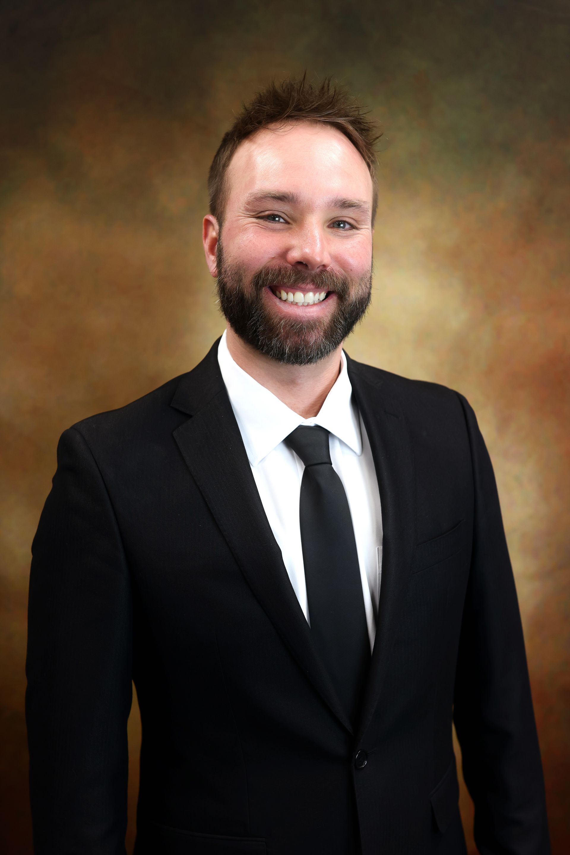 Smiling man in a black suit and tie against a warm studio backdrop