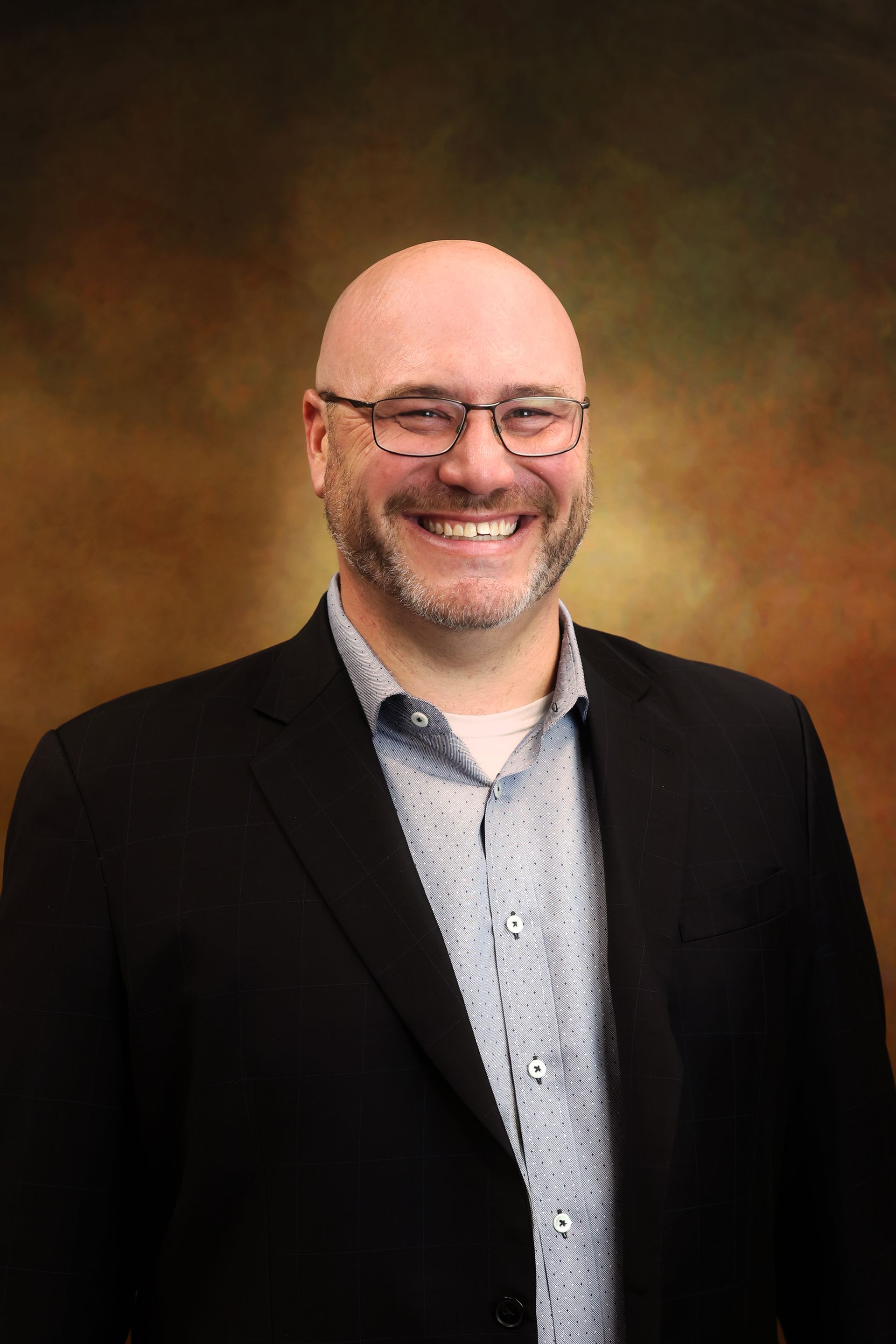 Smiling bald man in glasses wearing a dark blazer and patterned shirt against a warm brown backdrop