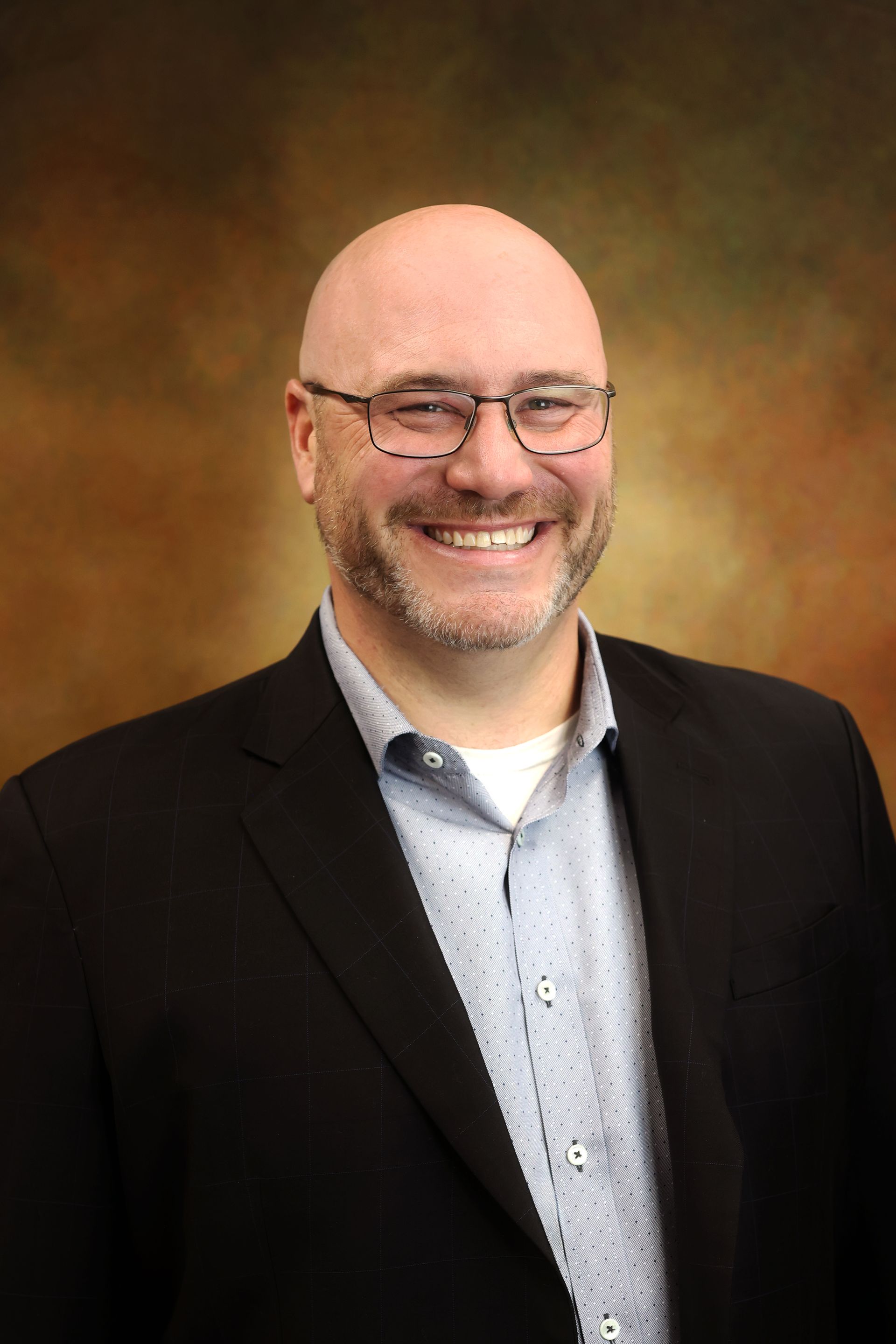 Smiling bald man in glasses and black blazer, posed against a warm studio backdrop