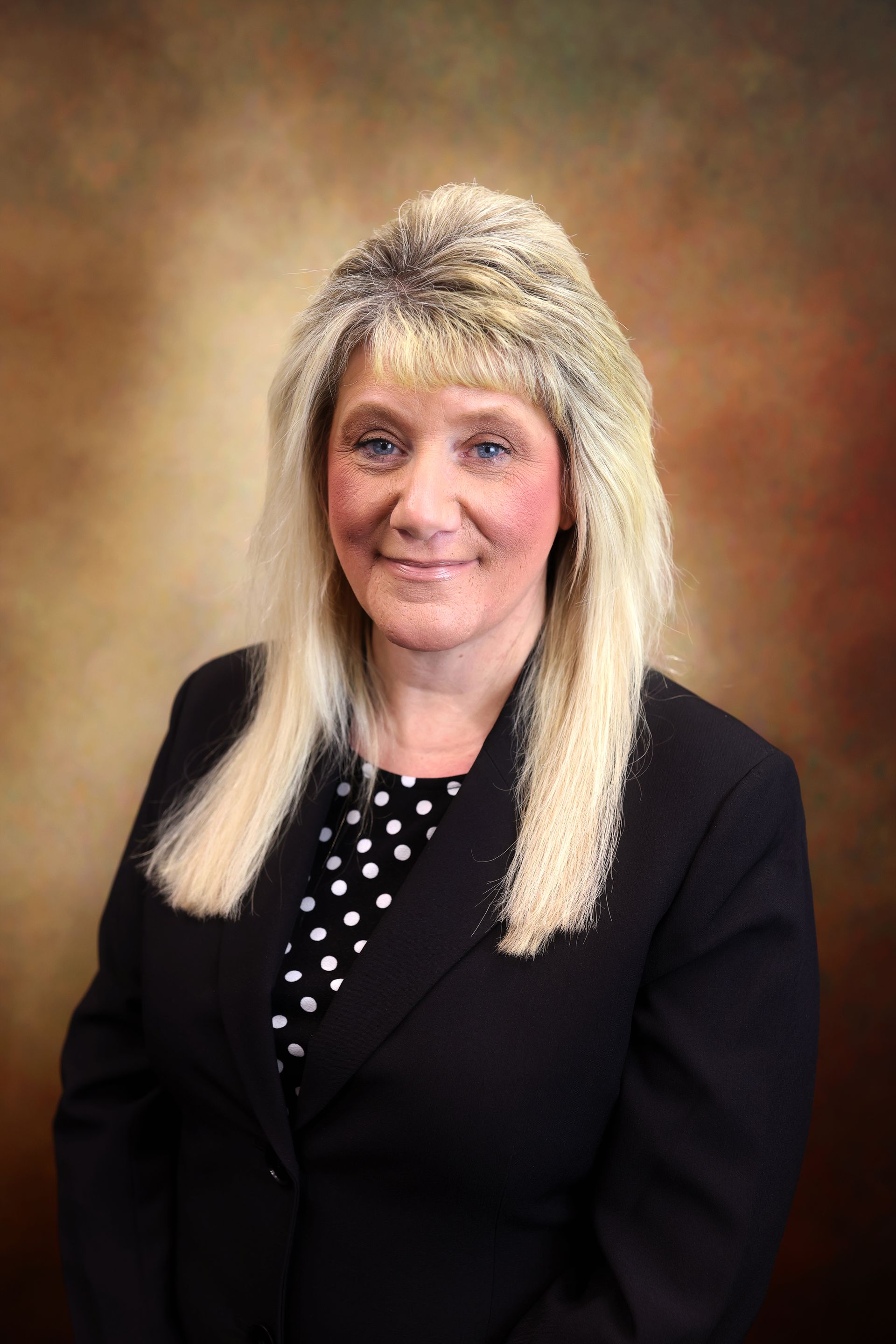 Portrait of a woman in a black blazer and polka-dot blouse against a warm brown backdrop