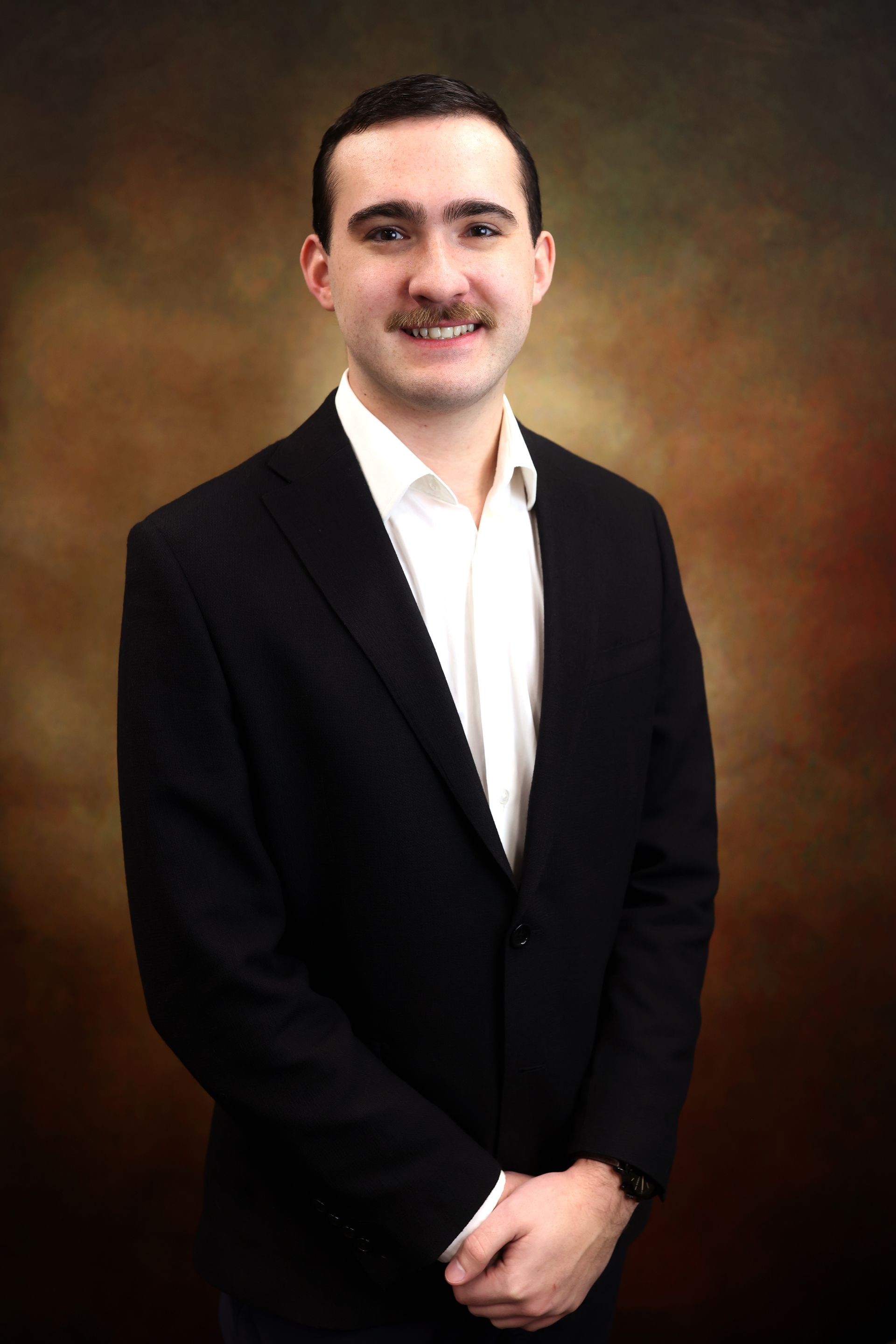 Studio portrait of a man in a black suit jacket and white shirt, standing against a brown backdrop.