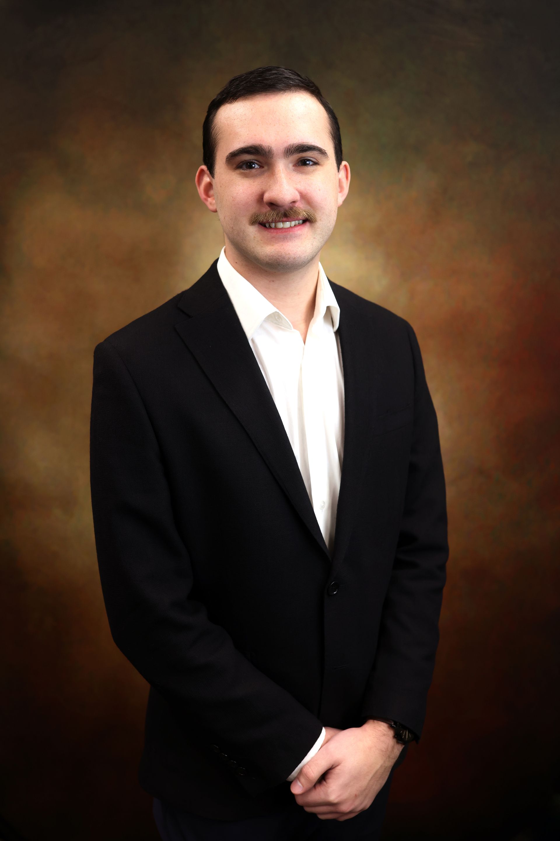 Man in a black suit and white shirt standing against a warm brown studio backdrop