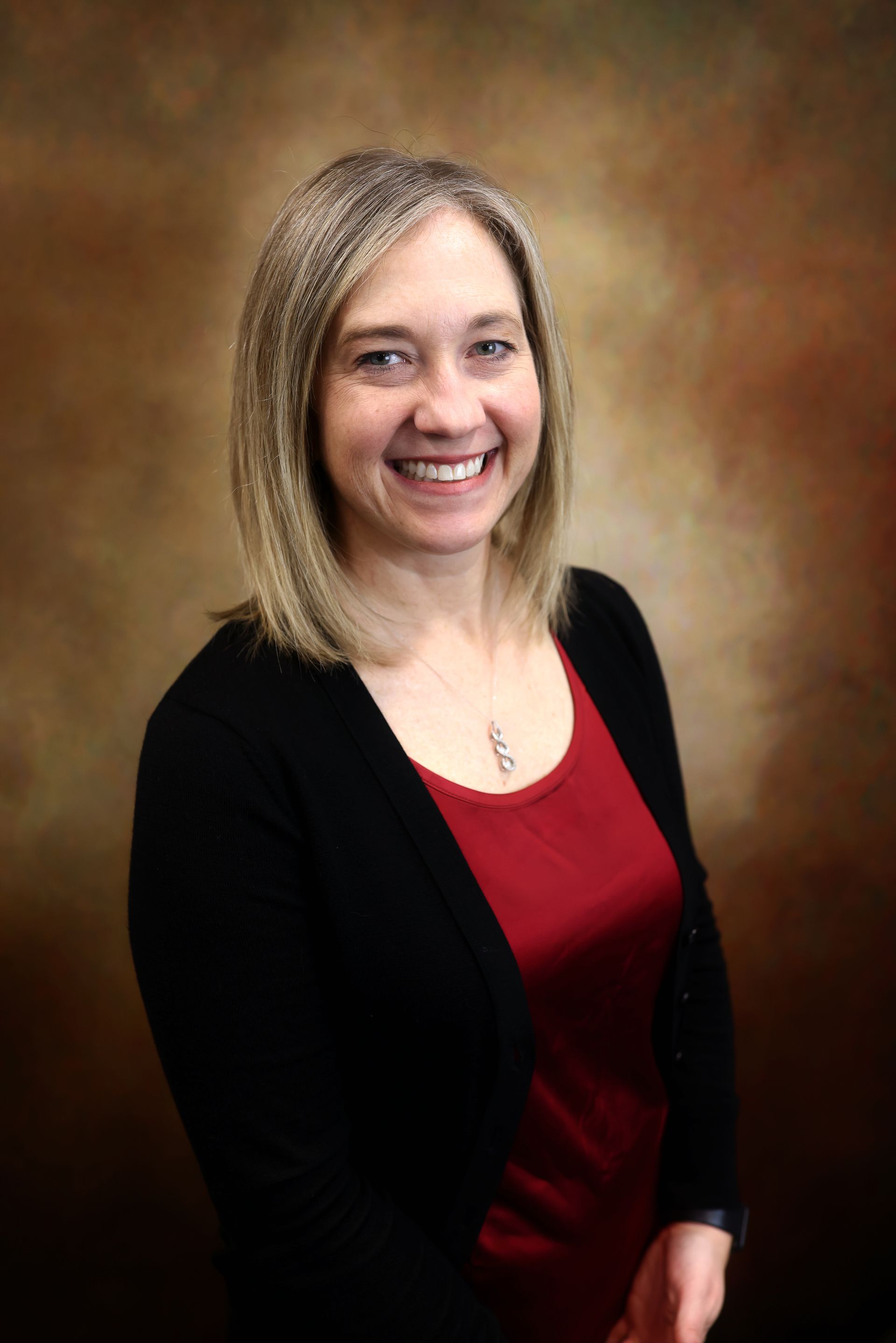 Smiling woman in a black cardigan over a red top, posed against a brown studio backdrop