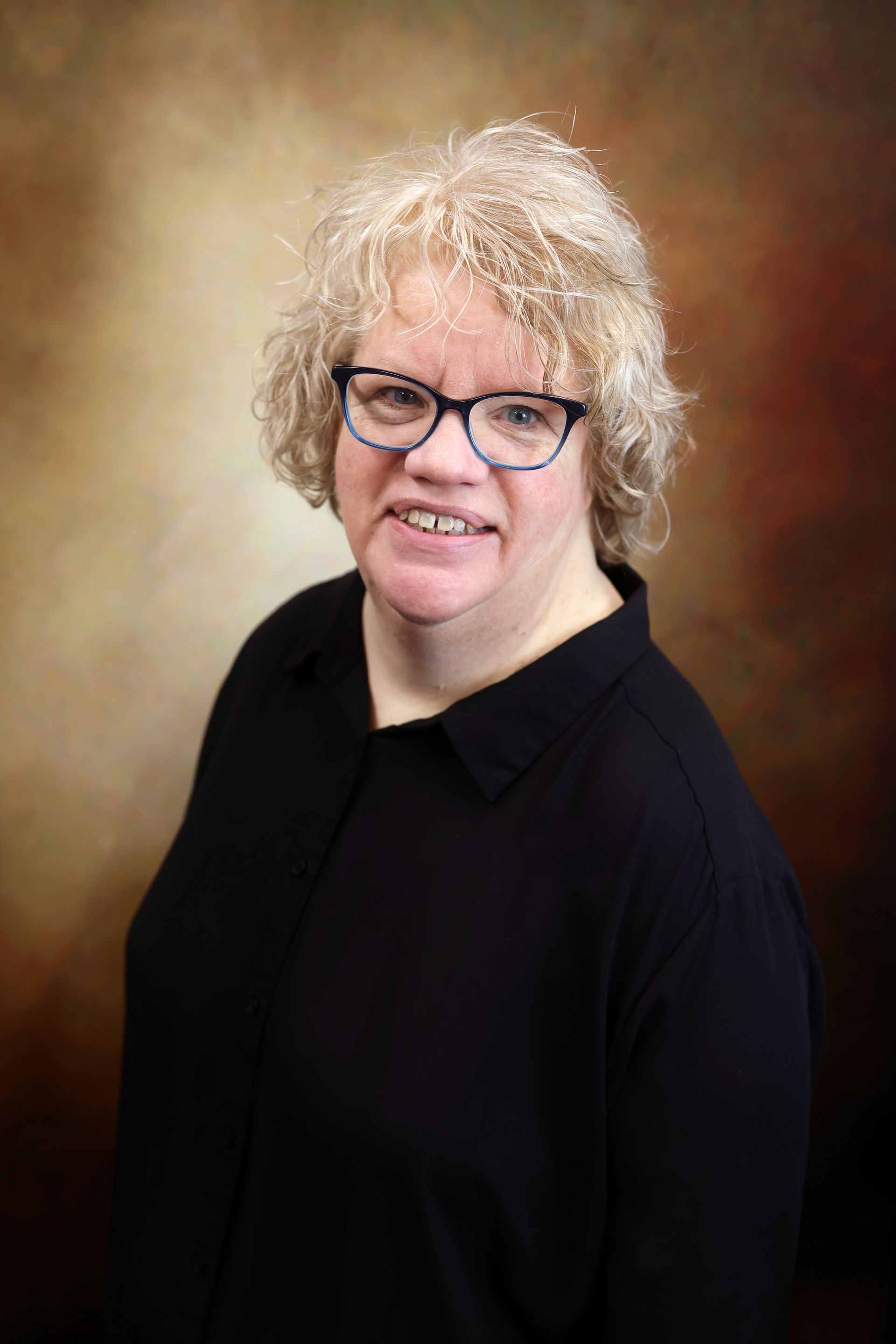 Smiling person with curly blond hair and glasses in a black top against a brown studio backdrop