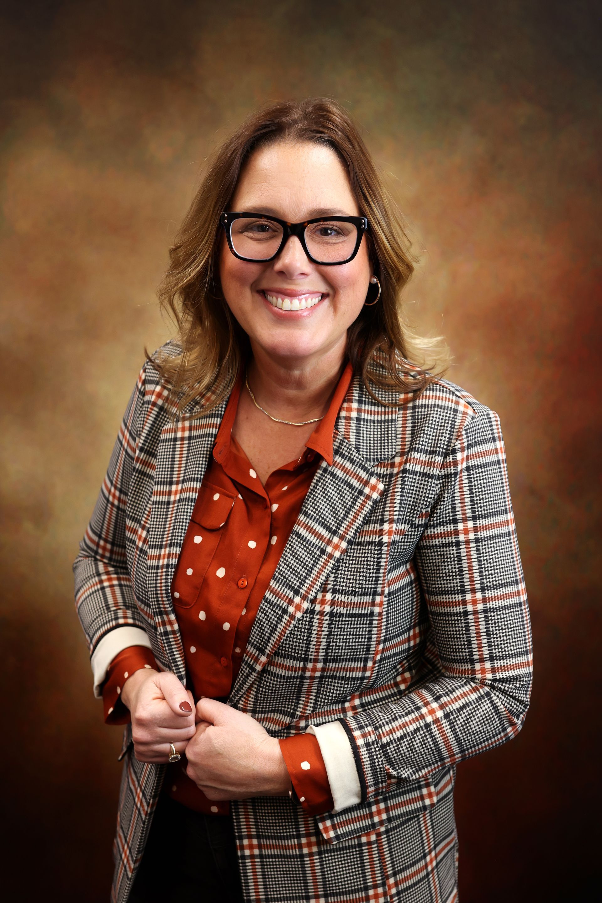 Smiling person in glasses wearing a plaid blazer and rust blouse against a brown studio backdrop