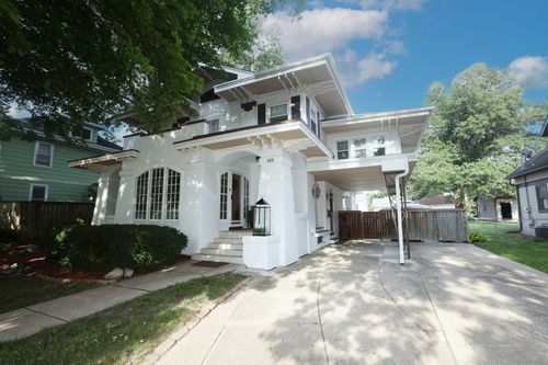 White two-story house with arched porch, large windows, and driveway under trees on a sunny day