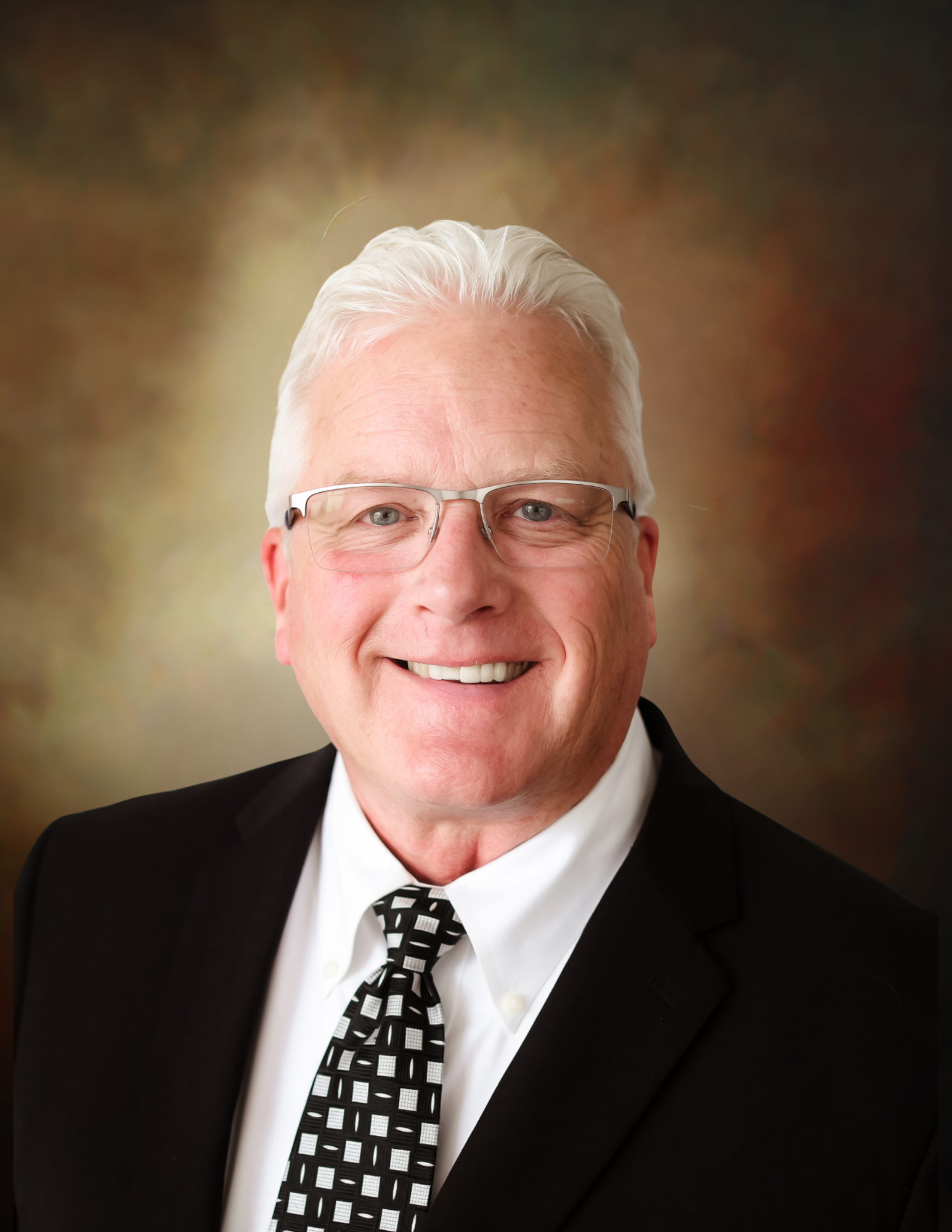 Smiling portrait of a man in a black suit, white shirt, and patterned tie against a blurred brown background