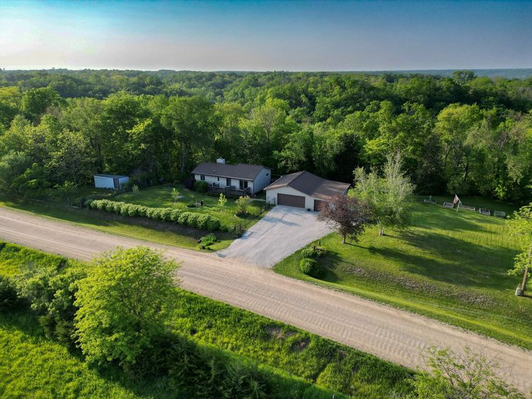 Aerial view of a rural house with a driveway beside a road, surrounded by green trees and fields