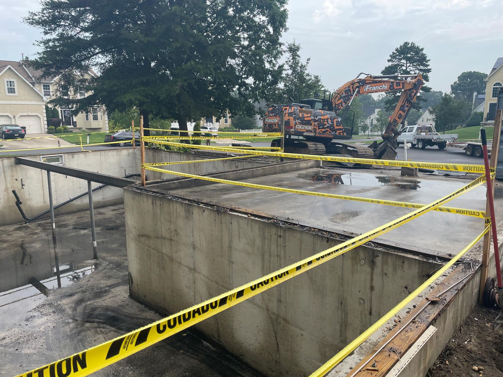 Construction site with concrete foundations, excavator, and caution tape. Residential neighborhood backdrop.