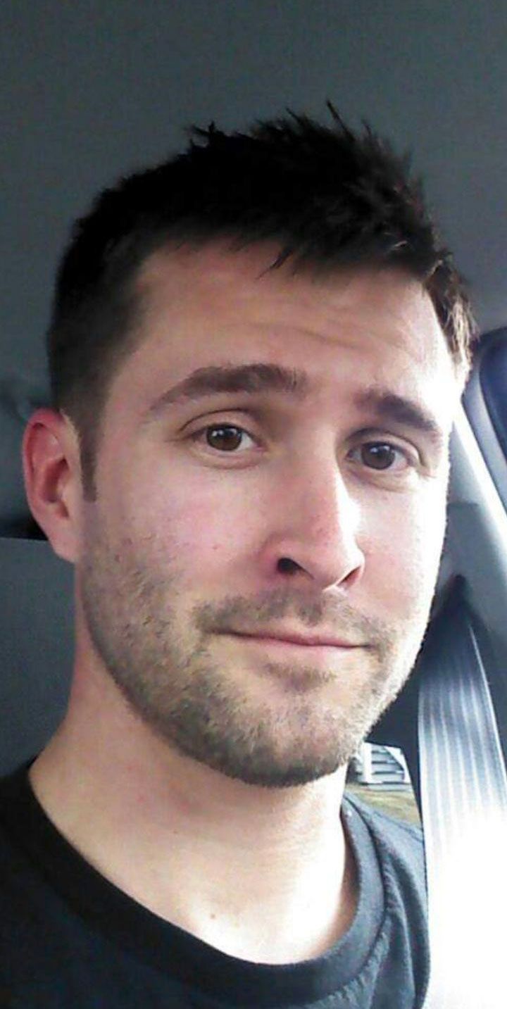 A portrait of a man named Kevin, identified as the Lead Commercial Electrician for Bee Electric, sitting in a vehicle. He has a short, styled haircut and light stubble. Kevin is wearing a black t-shirt and looking directly at the camera with a subtle smile, exuding a professional yet approachable demeanor.