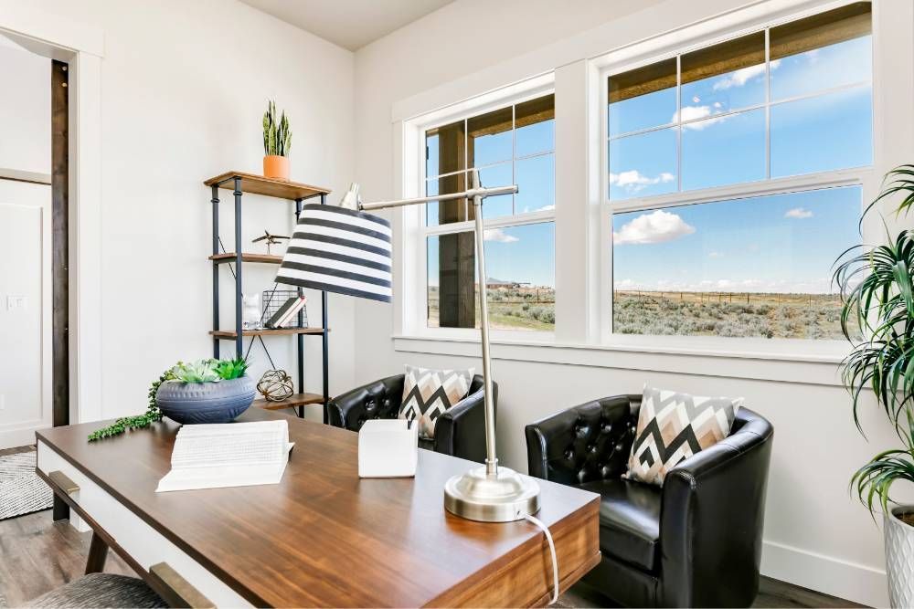 A home office with a desk , chairs , lamp and a large window.