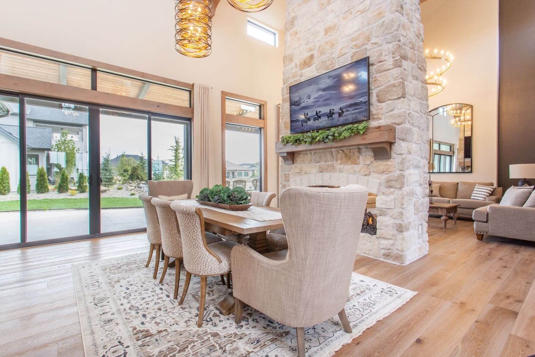 Elegant dining room with a large table, stone fireplace, and large windows overlooking a yard.