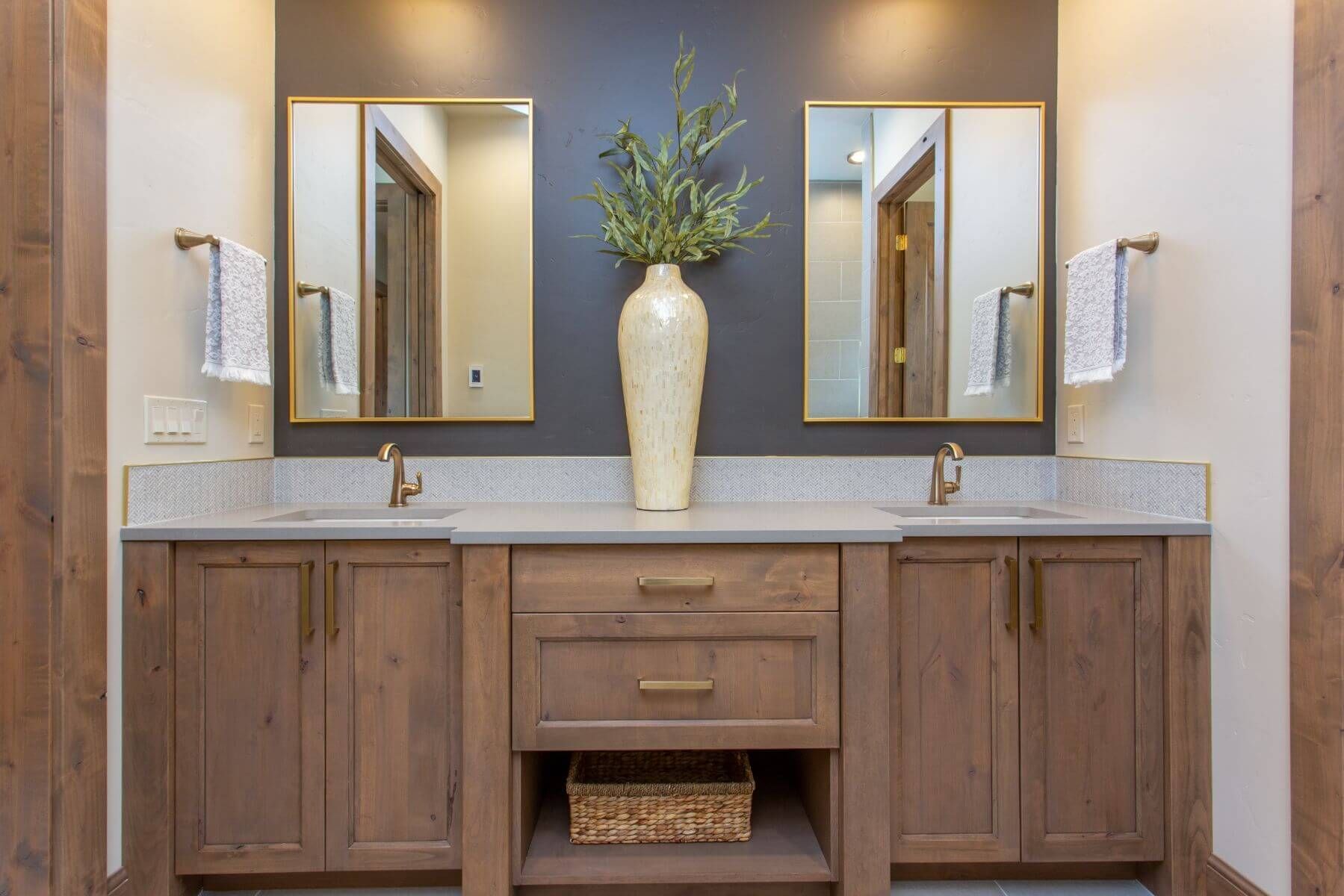 Bathroom with two sinks, wood cabinets, mirrors, and decorative vase against a dark gray wall.