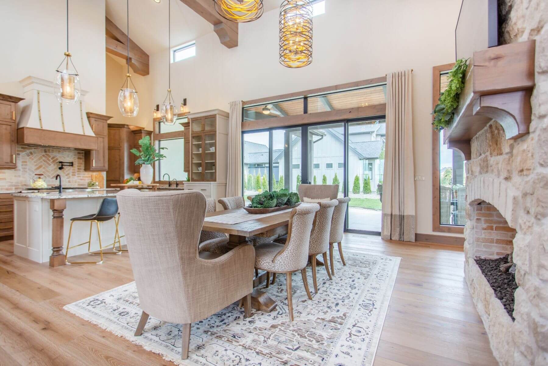 Open-concept kitchen/dining room with wood beams, stone fireplace, and large windows overlooking a yard.